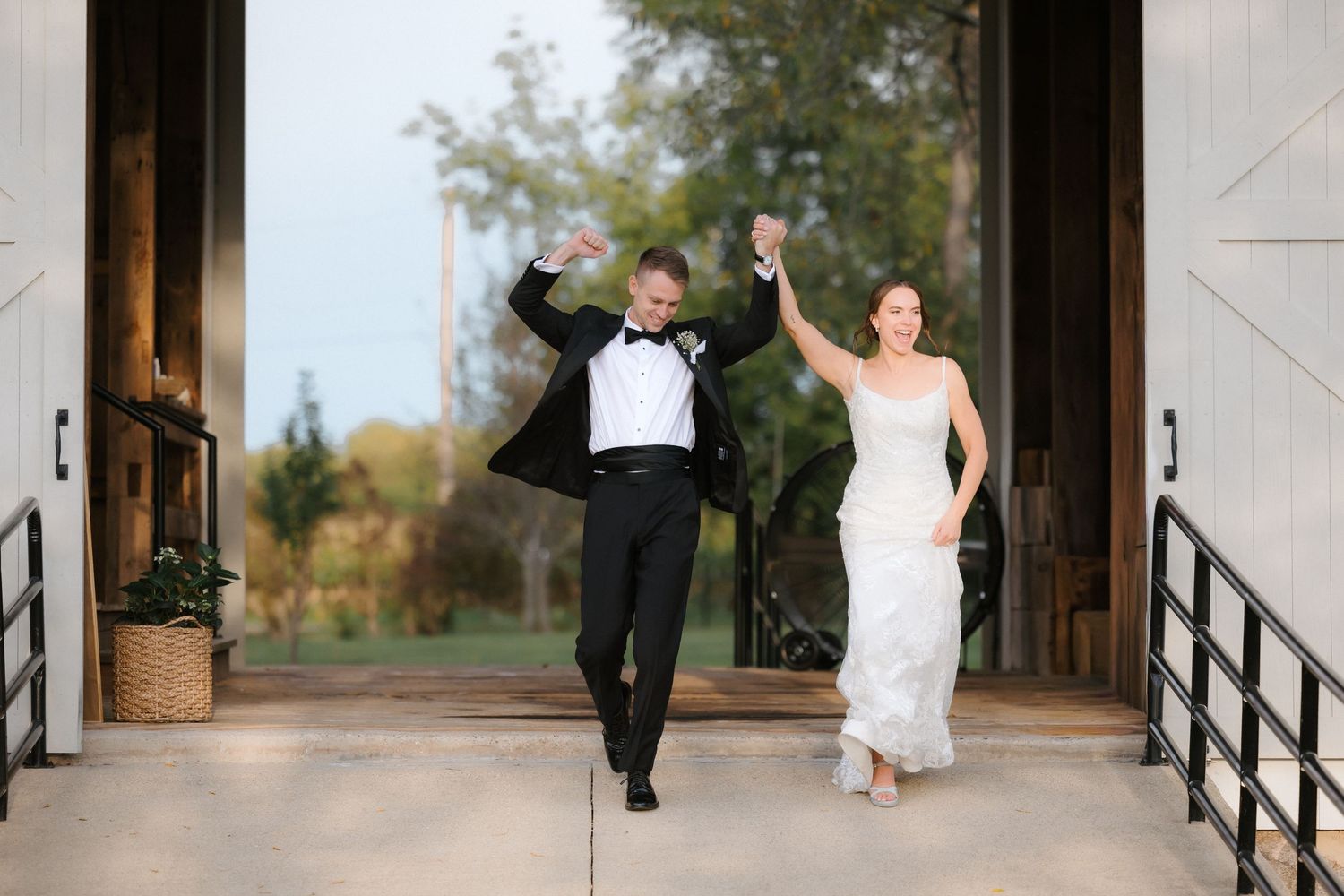 Joyful bride and groom raising arms in celebration as they exit through elegant white barn doors at their wedding reception.