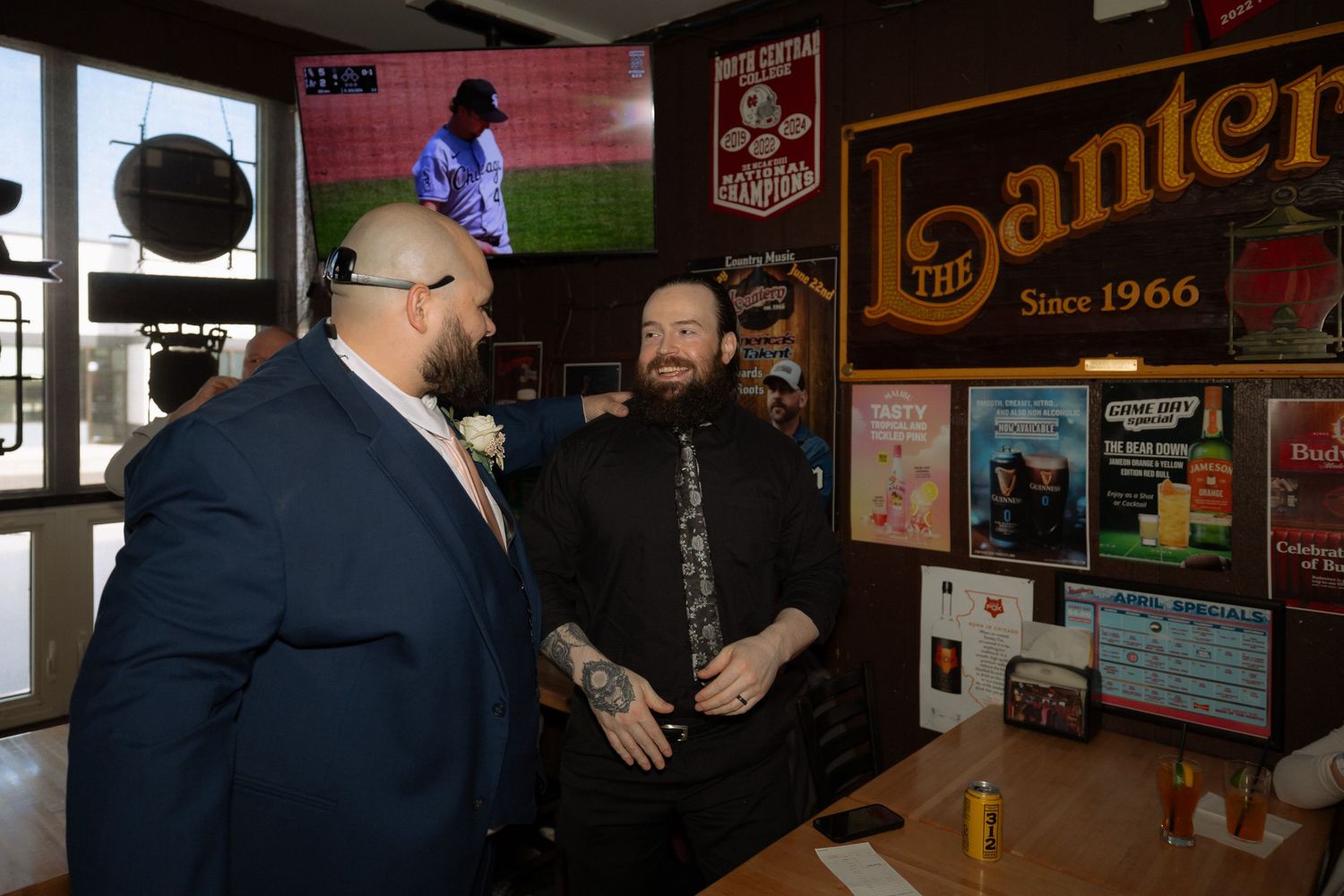 Two men in suits chat at The Lantern bar, established 1966, with sports TV and memorabilia in background.