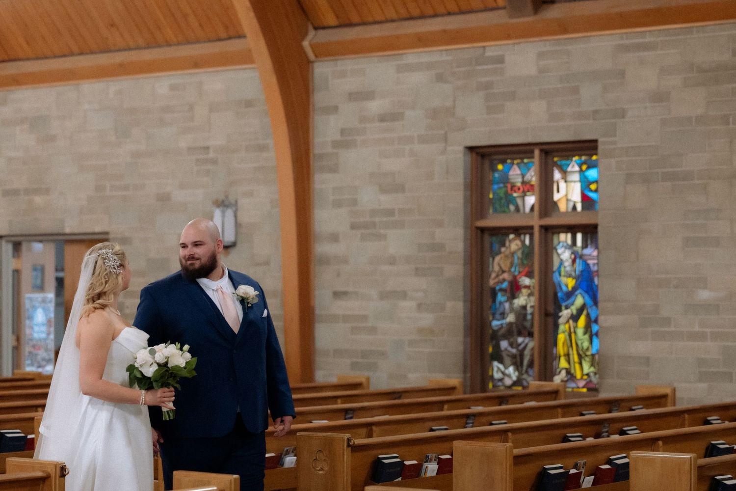 Bride and groom share a tender moment in a church aisle with stained glass windows and wooden pews.