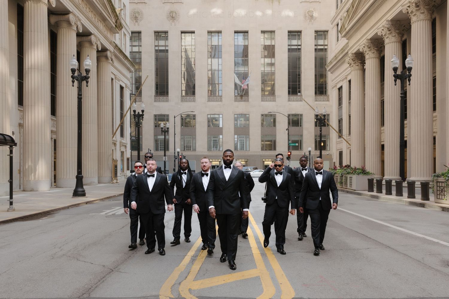 Groomsmen in black tuxedos and bow ties walk confidently down an urban street lined with classical columns.