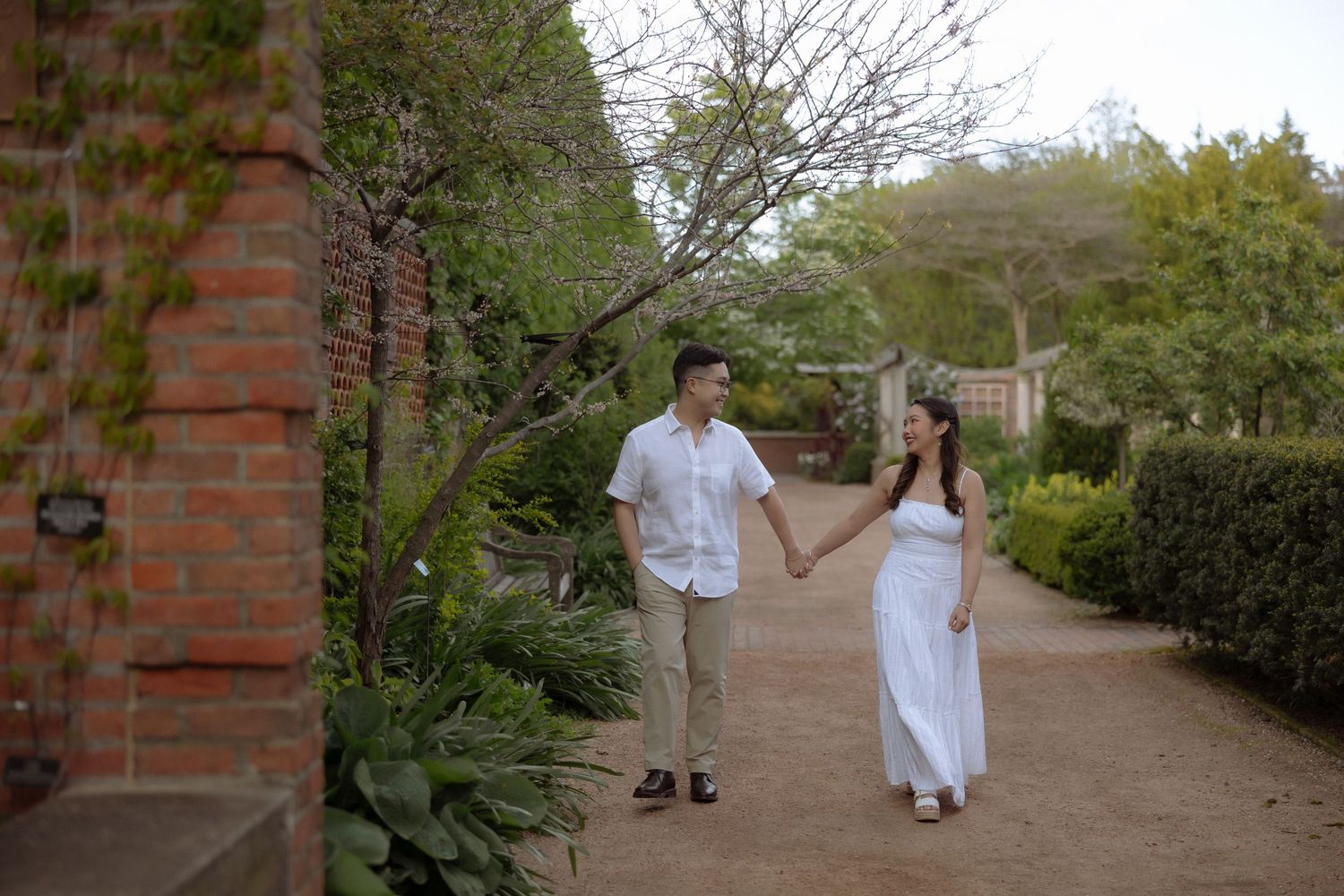 Couple holding hands walking along a garden path with brick walls and lush greenery in a romantic outdoor setting.