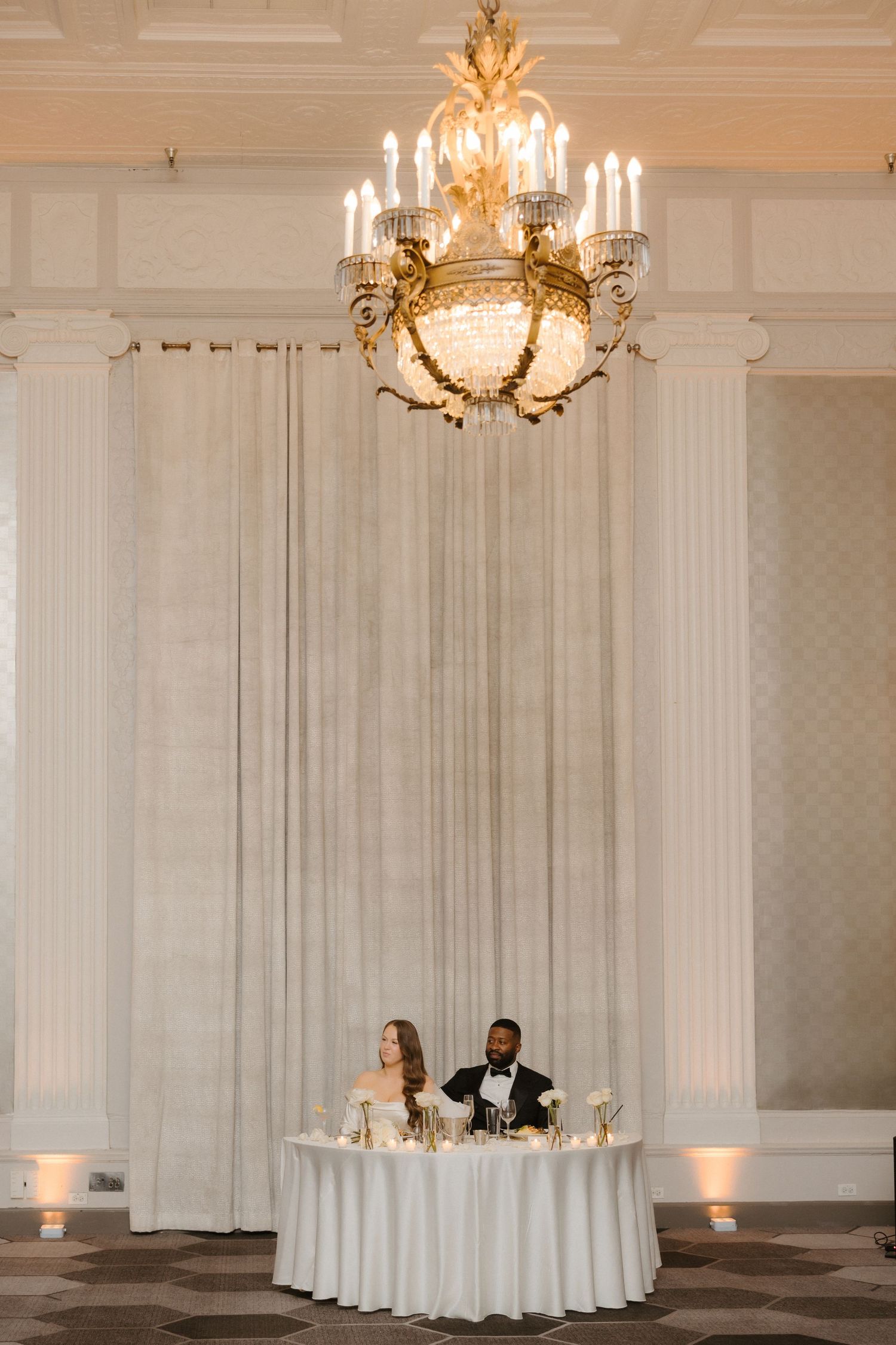 Bride and groom seated at sweetheart table beneath elegant crystal chandelier in grand ballroom wedding reception.