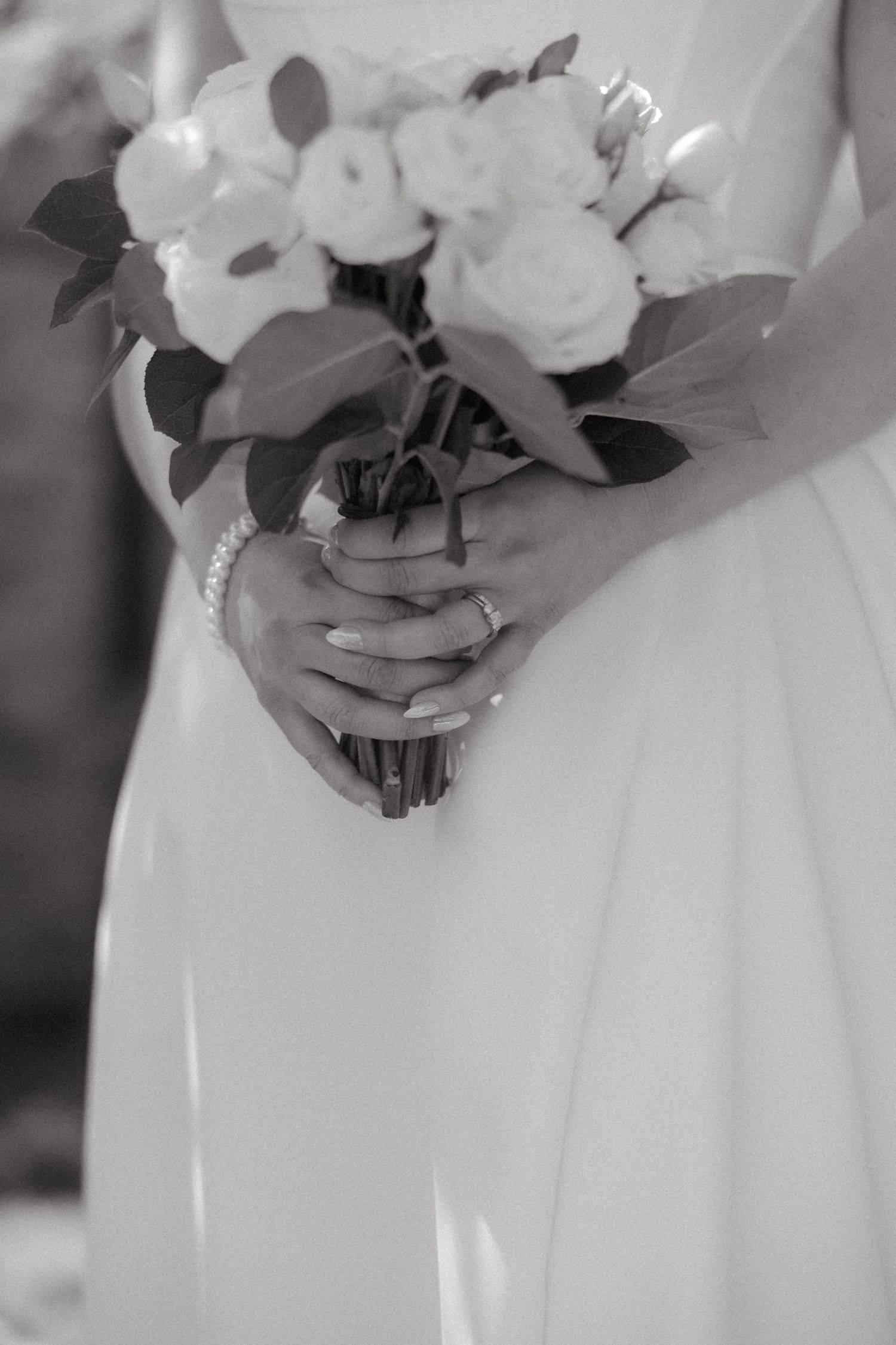 Black and white photo of a bride in white dress holding a bouquet of white roses with pearl bracelet and ring.