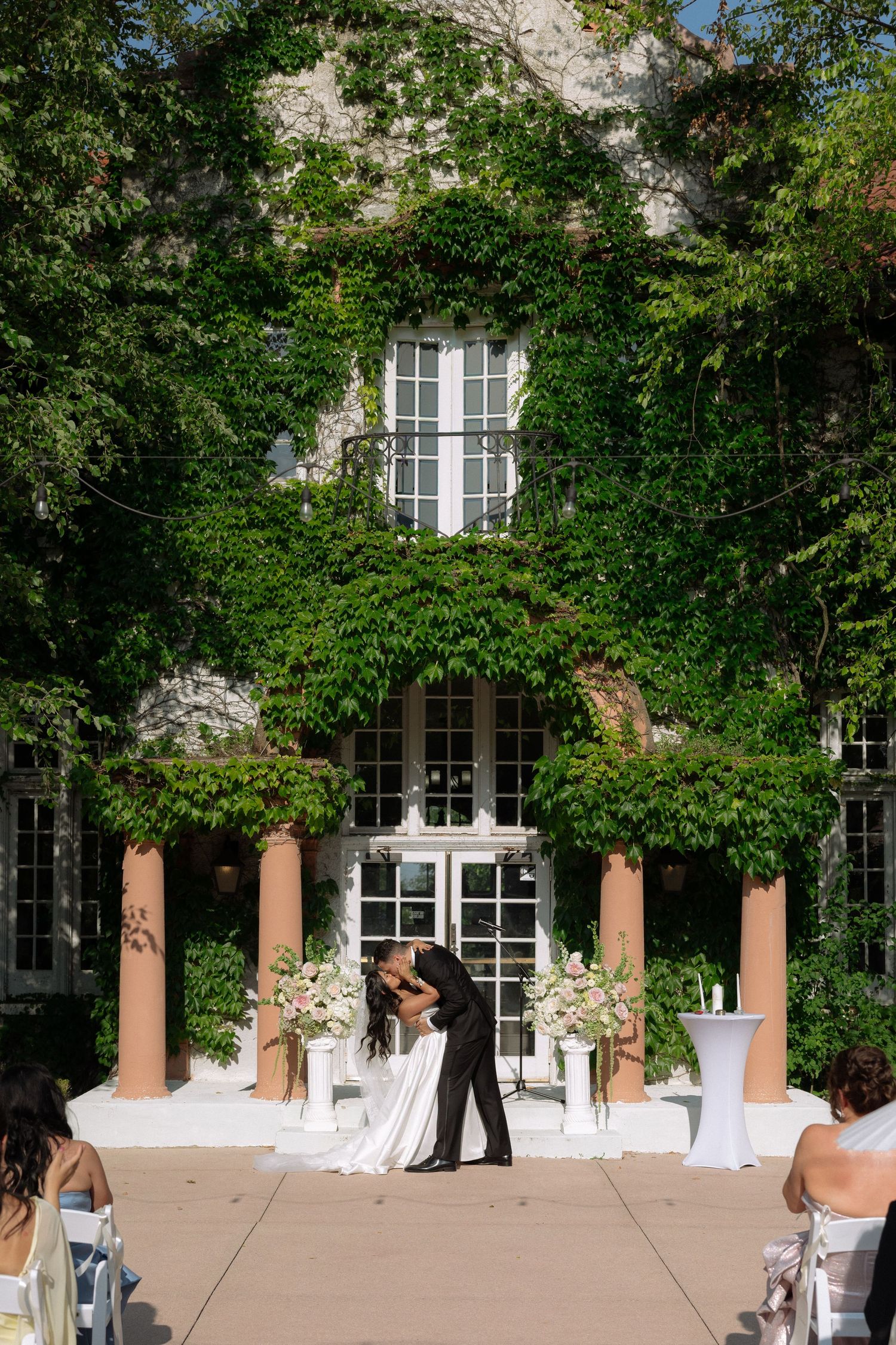 Bride and groom share their first kiss beneath an ivy-covered building flanked by white floral arrangements.
