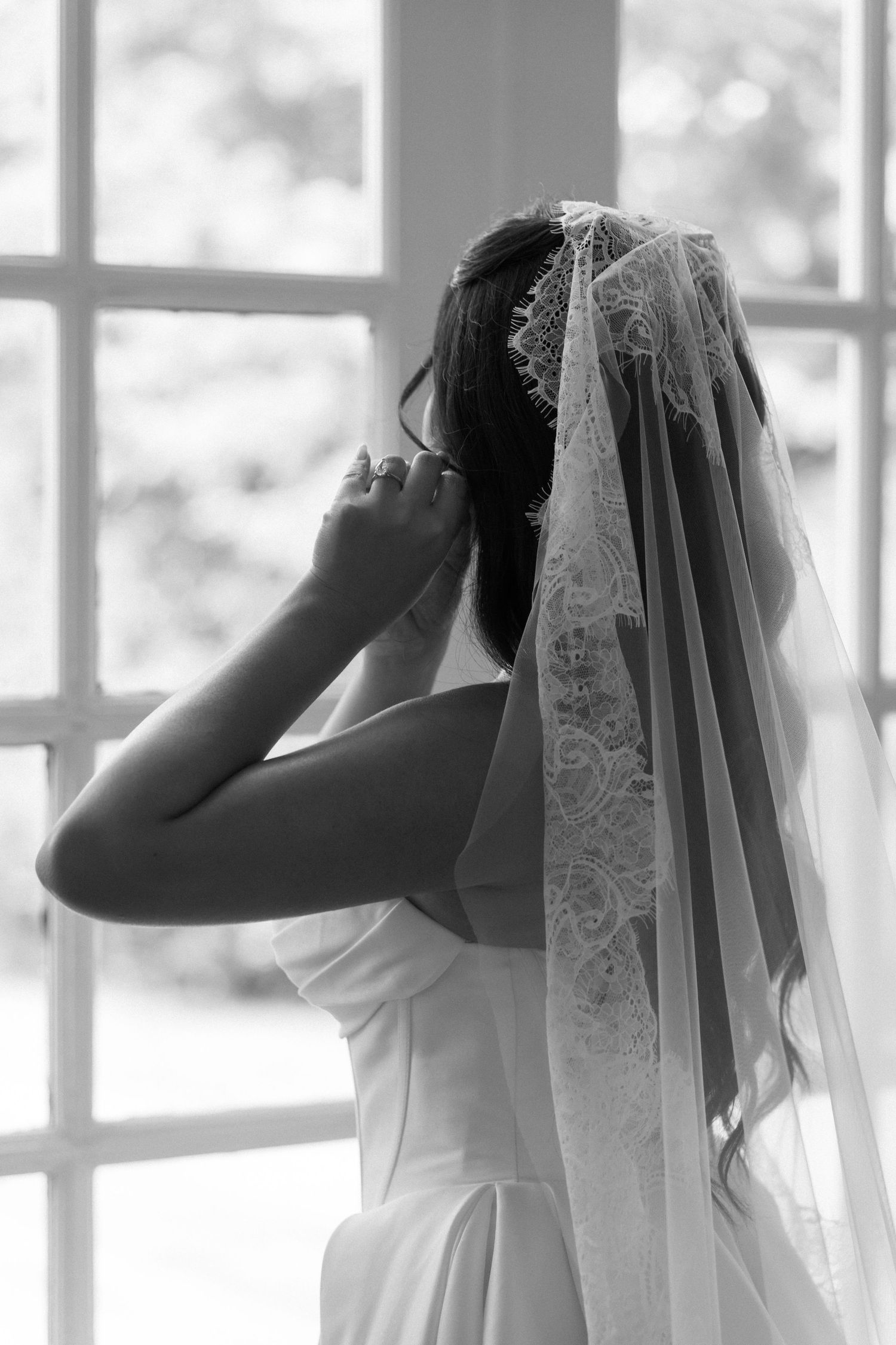 Bride in white gown and lace veil stands before window in black and white portrait.