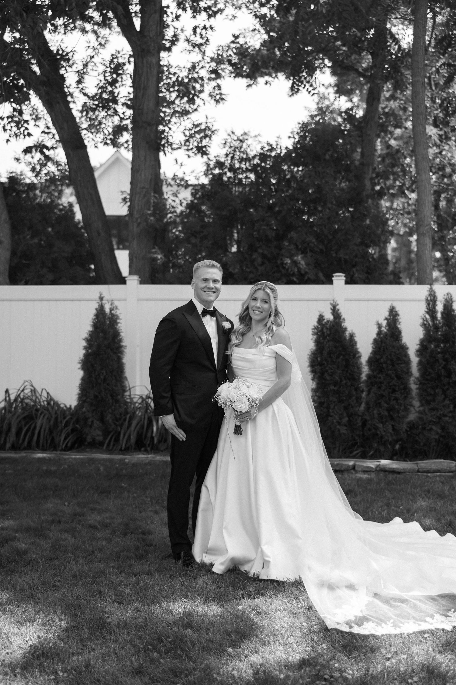 Bride and groom pose together in black and white portrait on lush lawn with evergreen trees.