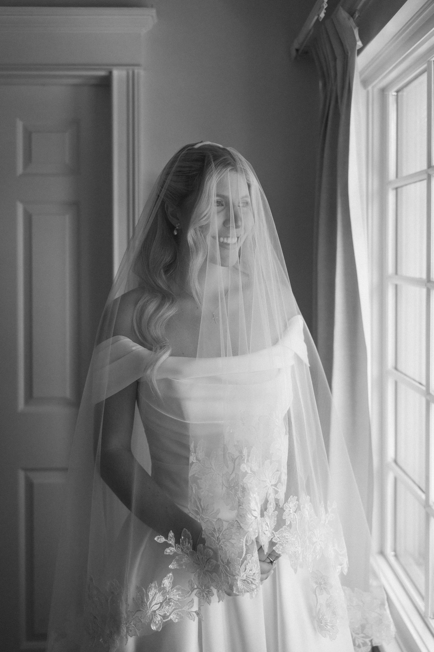 Smiling bride in white gown and veil holds bouquet by window in black and white wedding portrait.