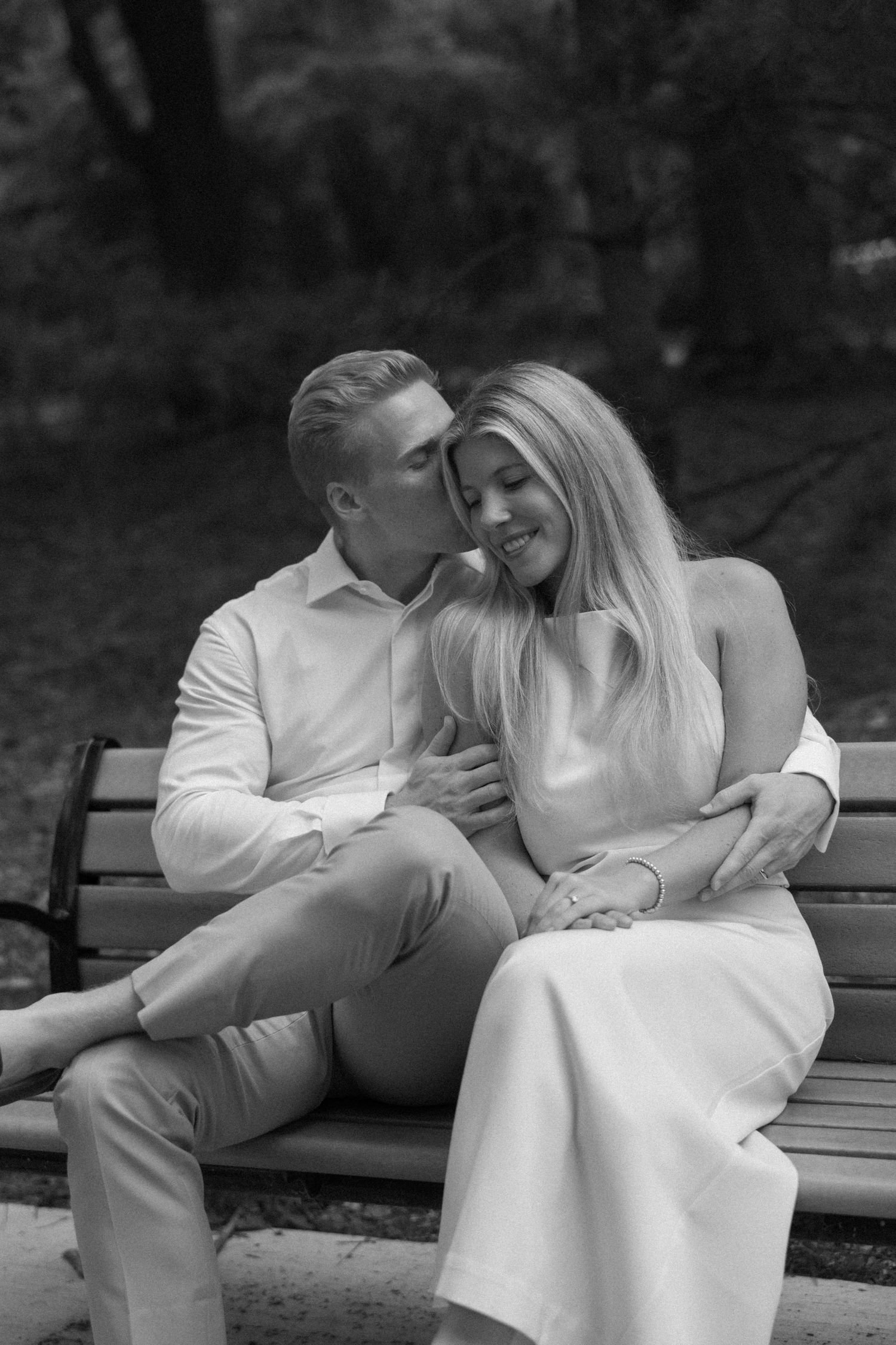 A couple shares a tender moment on a park bench, man kissing woman's cheek while embracing her in black and white.
