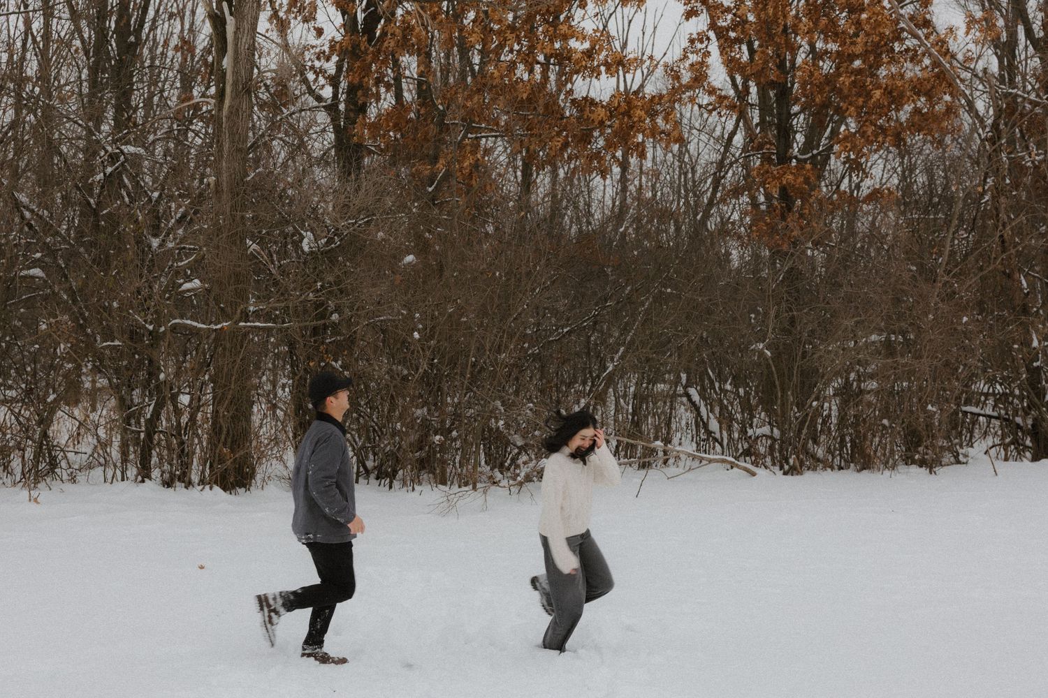 Two people walking through a snowy winter landscape with bare trees and autumn foliage in the background.