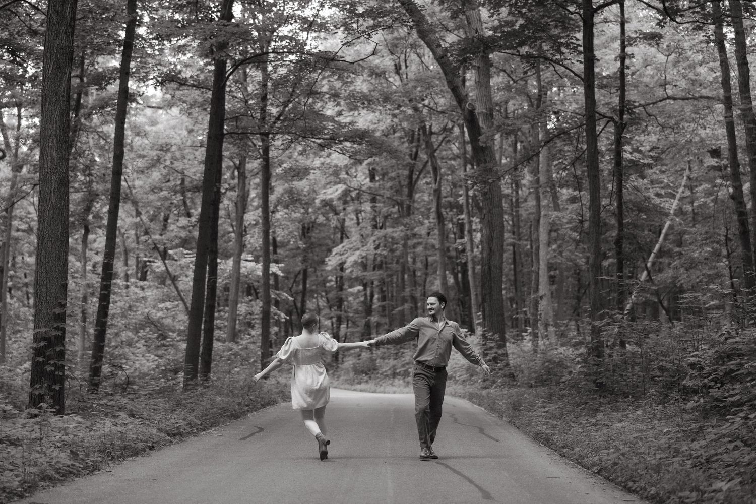 Black and white photo of a couple holding hands and running down a tree-lined road in a lush forest.