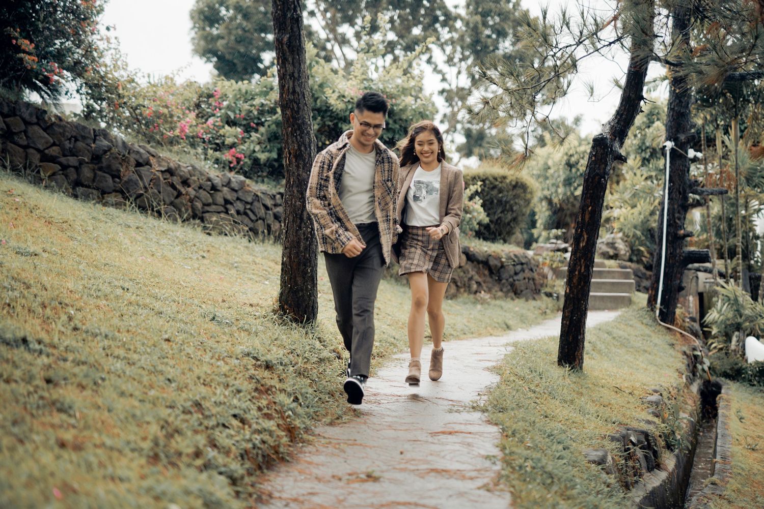 A couple walks hand in hand along a garden path surrounded by lush green trees and grass.
