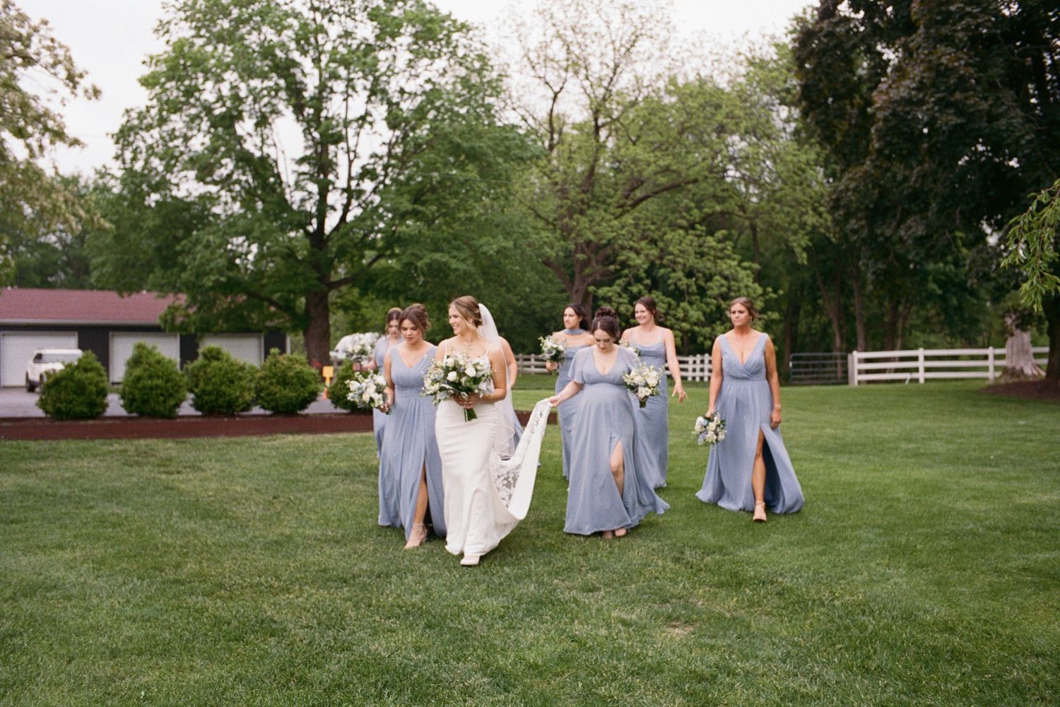 Bride in white gown walks with five bridesmaids in dusty blue dresses across a lush green lawn at an outdoor wedding.