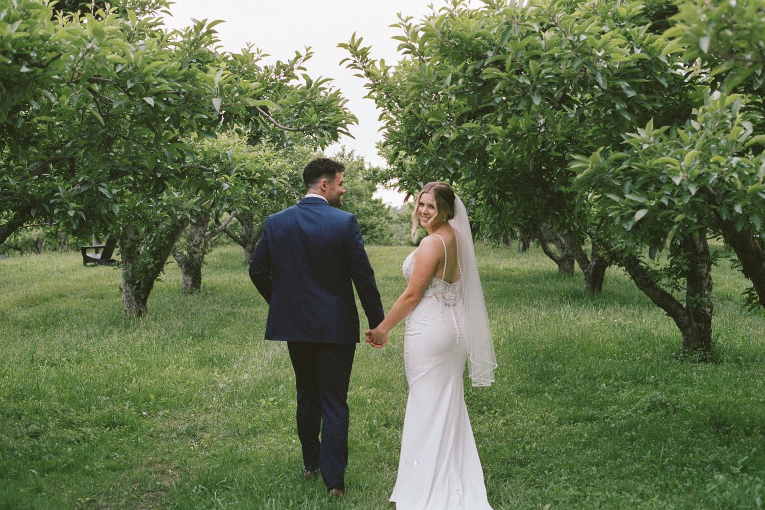 Bride and groom walk hand-in-hand through a lush green orchard on their wedding day.