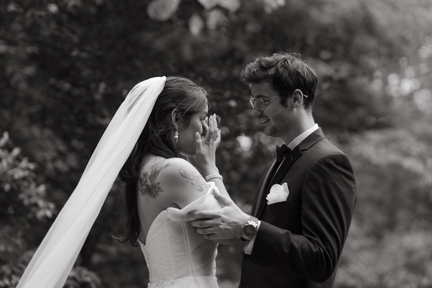 Black and white photo of emotional bride wiping tears while groom smiles during first look.