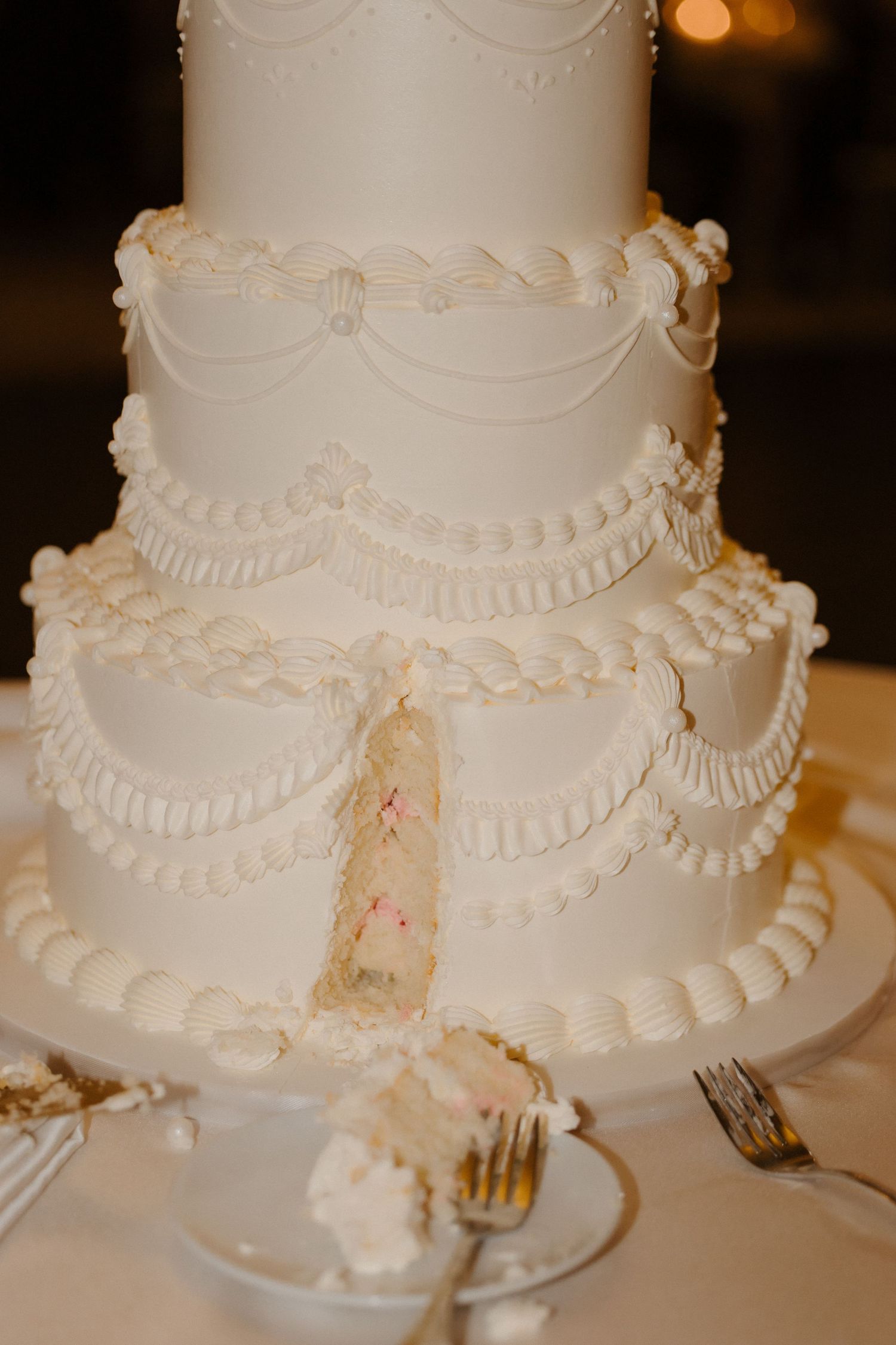 Elegant three-tier white wedding cake with intricate piped frosting details and draped swag decorations, with a slice cut out.