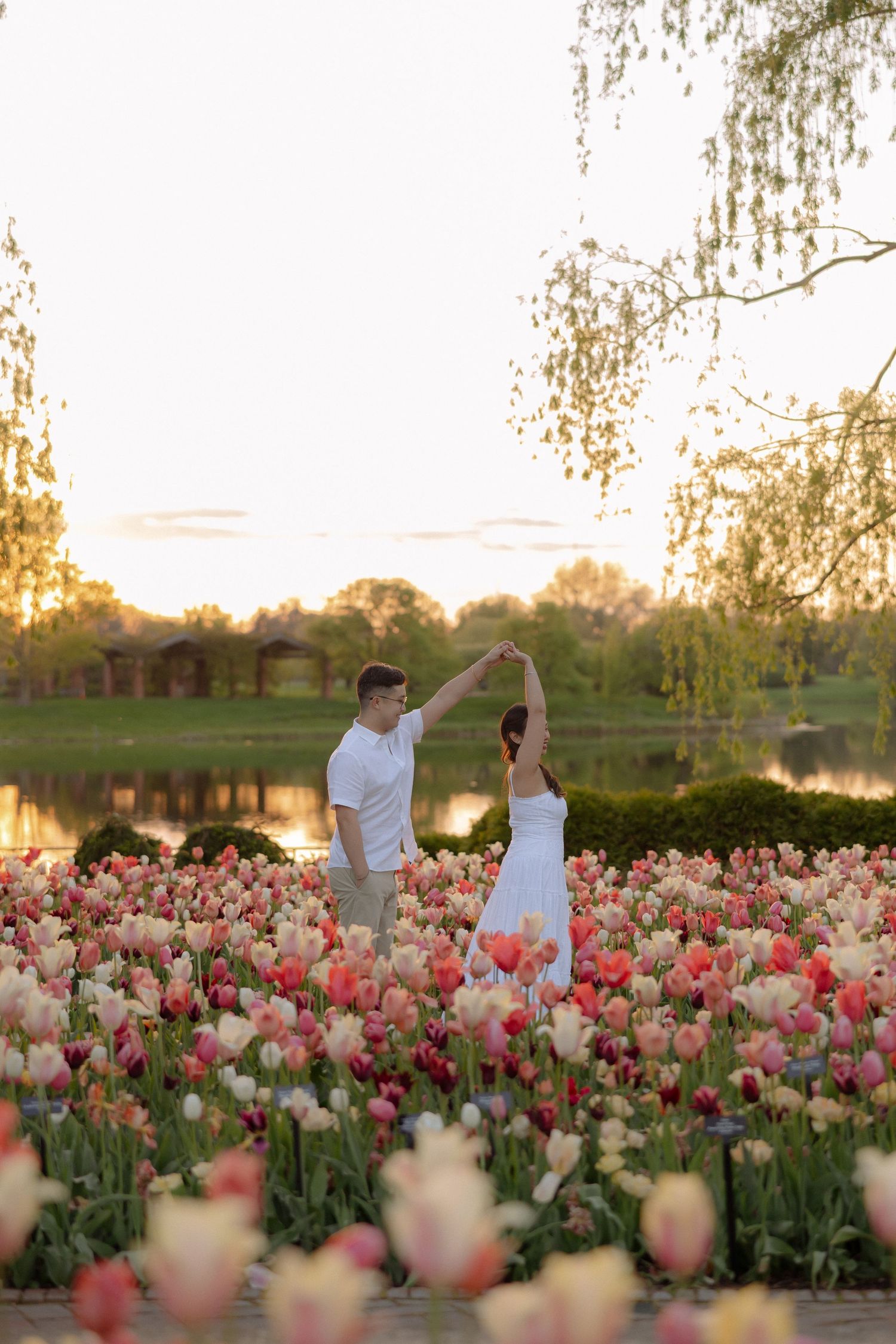 Couple dancing in a vibrant tulip field at sunset near a serene lake, surrounded by pink and red blooms.