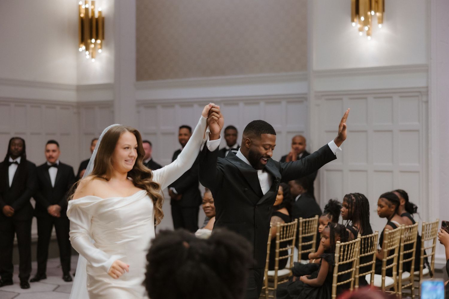 Bride and groom joyfully enter wedding reception, bride in off-shoulder gown, groom in black tuxedo, elegant ballroom.