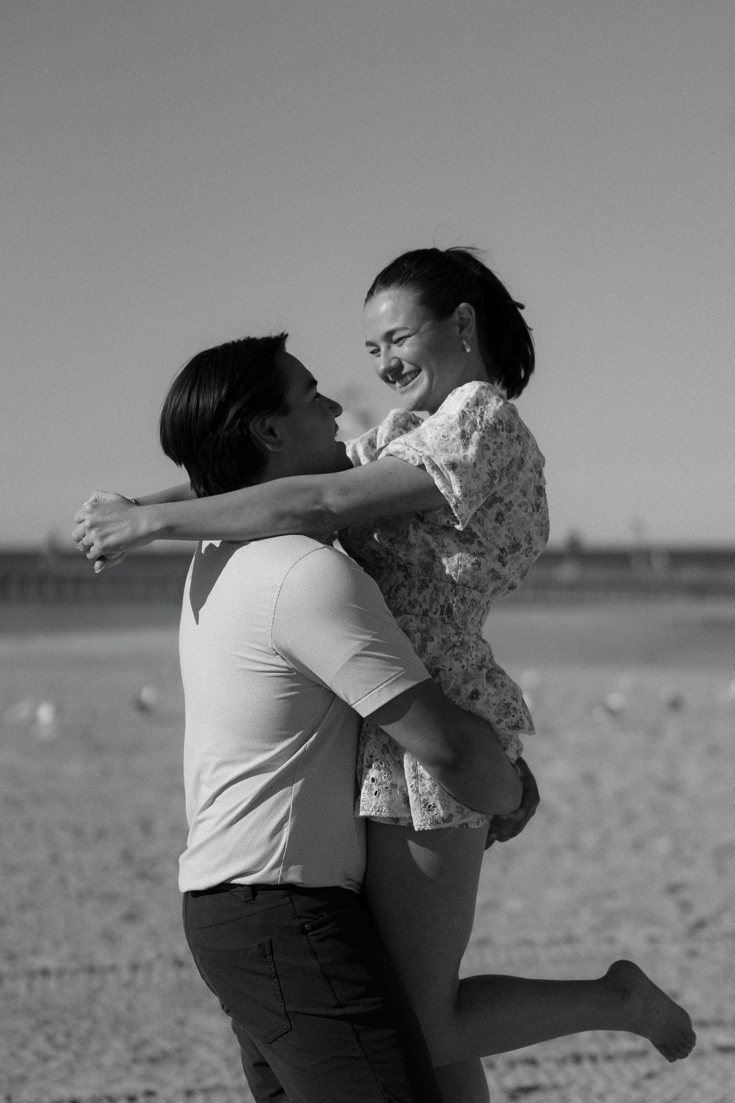 Black and white photo of man lifting smiling woman on beach during romantic outdoor session.