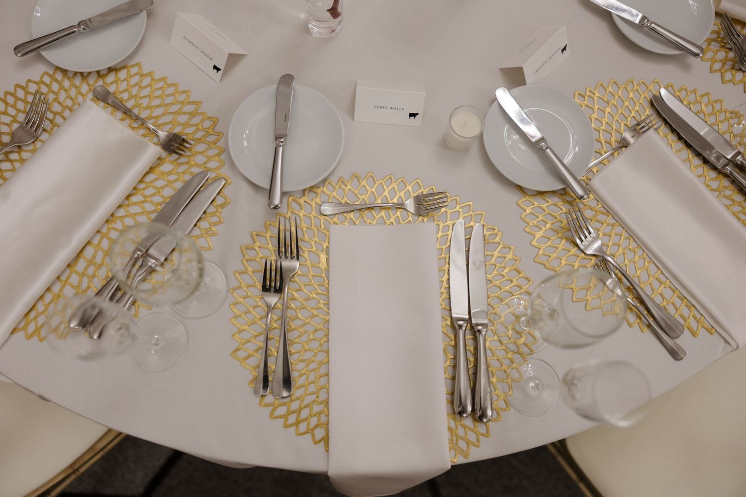 Elegant round dining table set with gold placemats, silver cutlery, white napkins, and crystal glasses for a formal event.