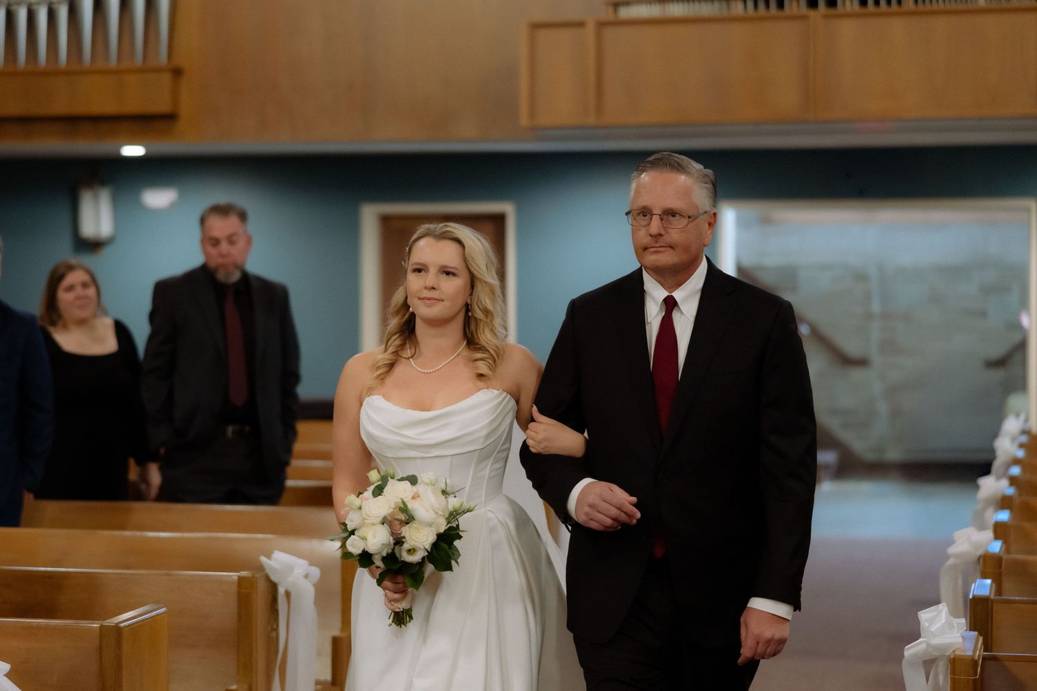 Bride in white gown walks down church aisle with father, holding white bouquet during wedding ceremony.