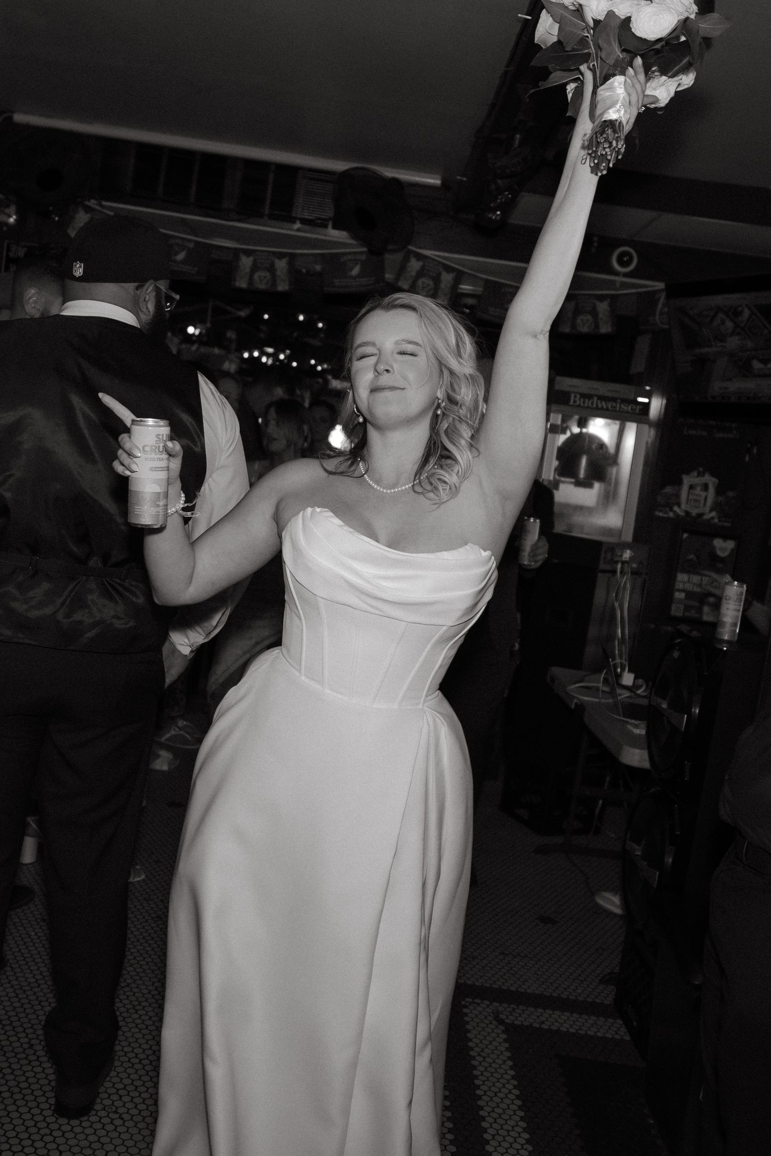 Bride in strapless gown raises bouquet triumphantly while holding drink at wedding reception in black and white photo.