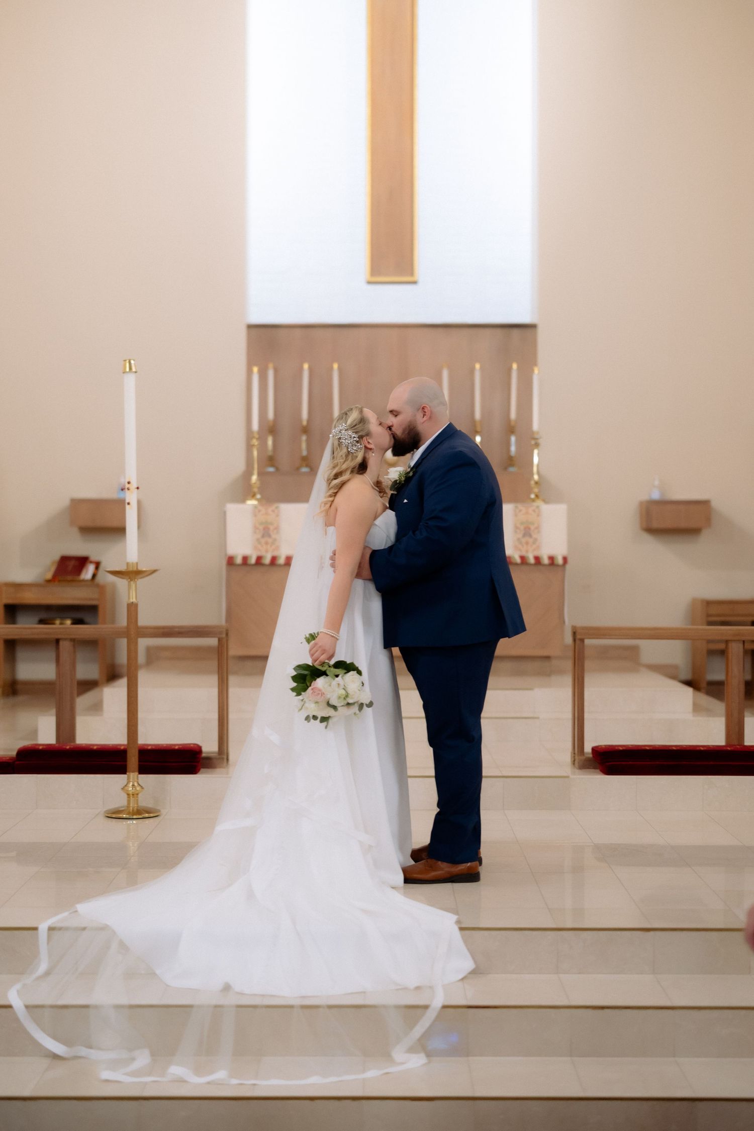 Bride and groom share a kiss at the altar of a church, bride holding a bouquet in a white gown with long veil.