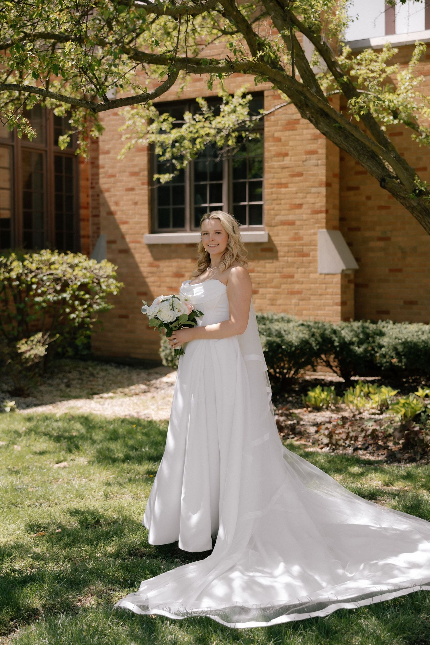 Smiling bride in white gown with train holds bouquet outdoors by brick building and green trees on sunny day.