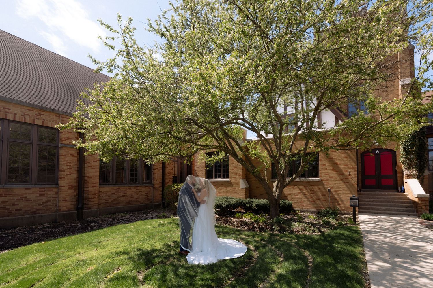 Bride in white gown and veil stands beneath a blooming tree outside a brick church with red doors on a sunny day.