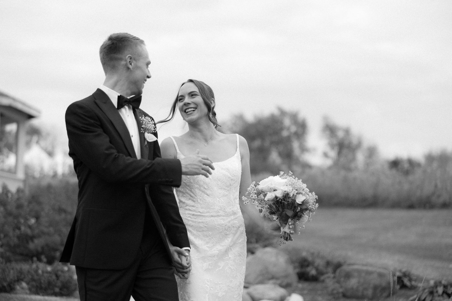 Black and white portrait of smiling bride holding bouquet walking with groom outdoors.