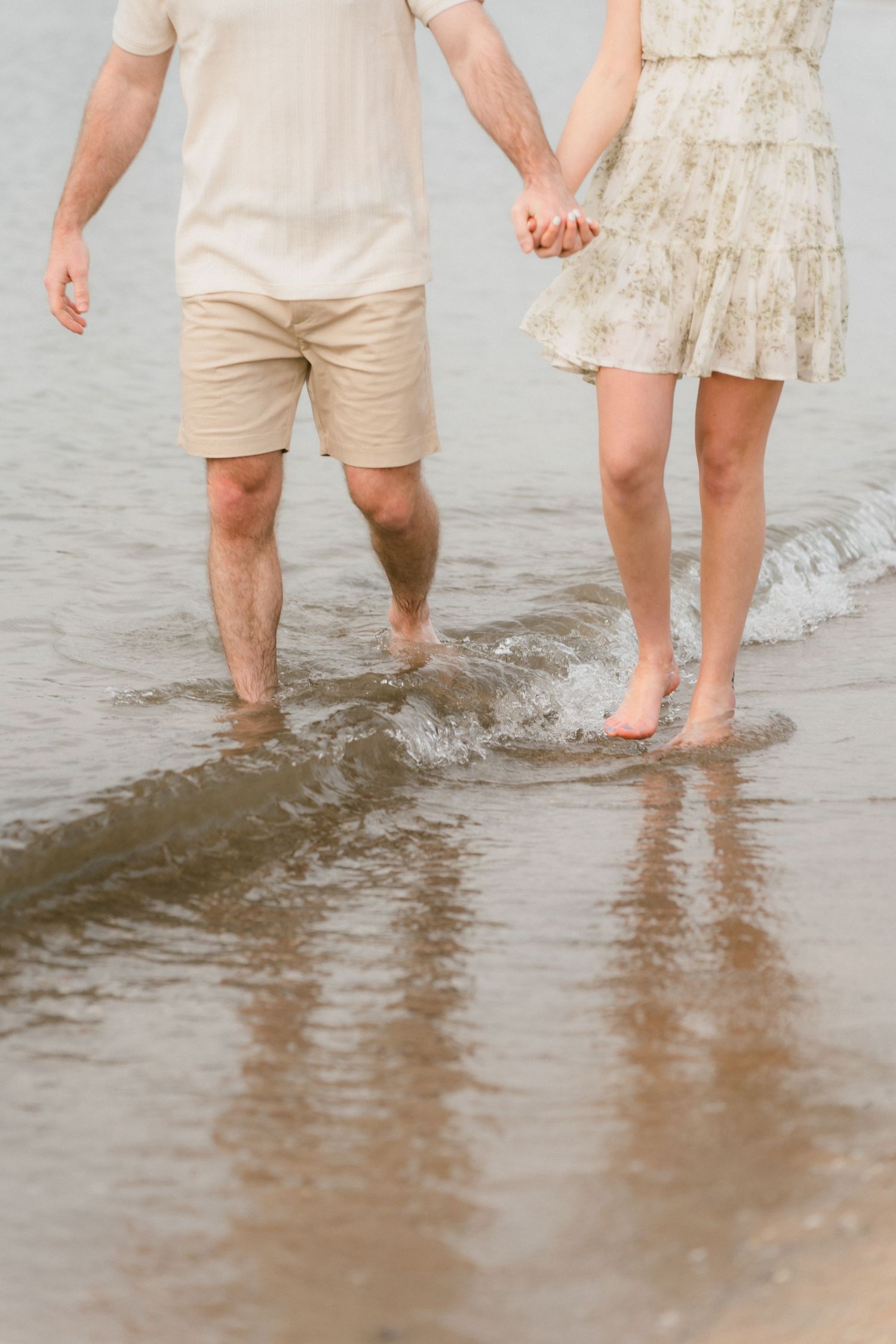 Barefoot couple holding hands while wading in shallow water along a sandy shoreline.