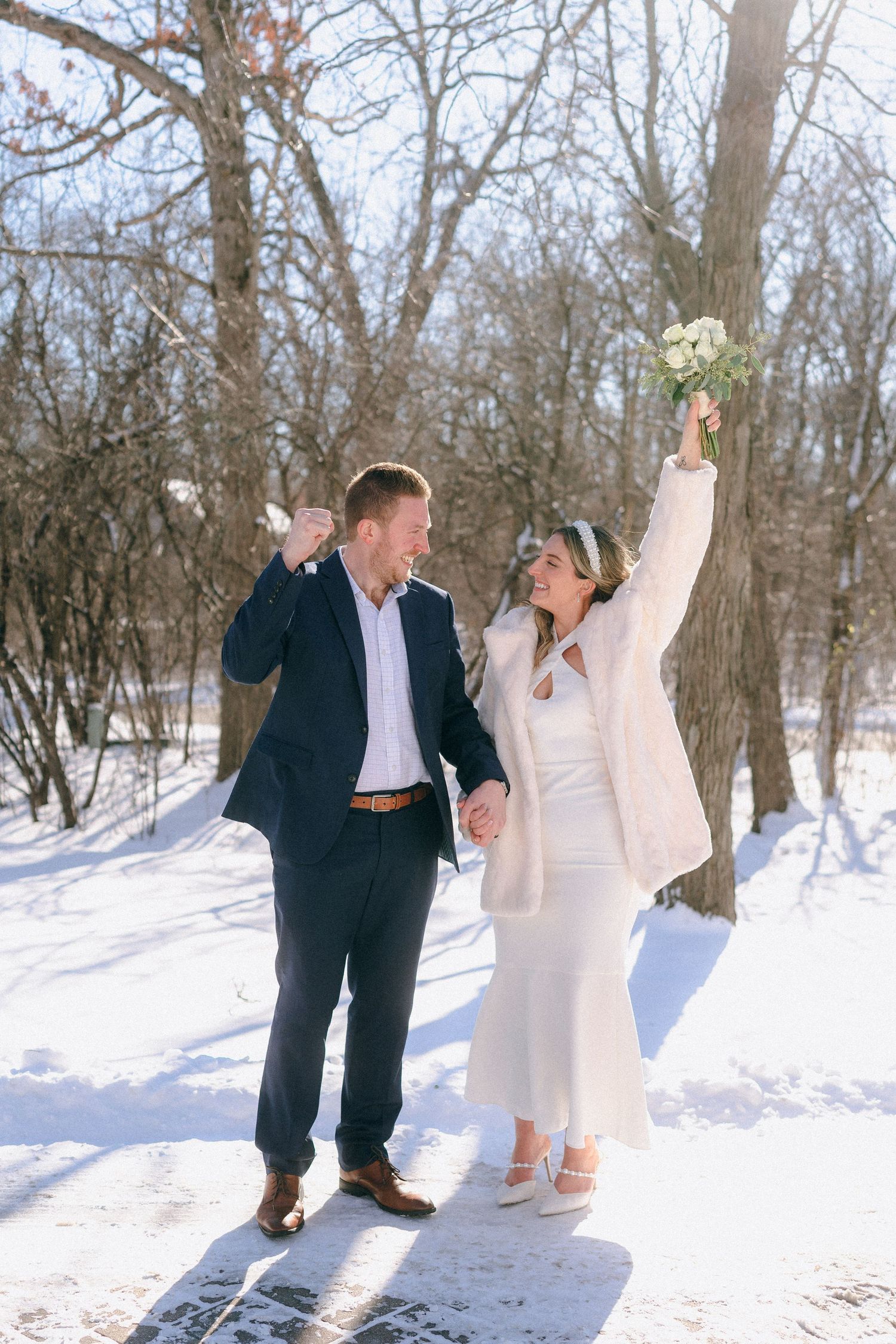 Joyful newlywed couple celebrating in a snowy winter wonderland, bride raising bouquet in the air.