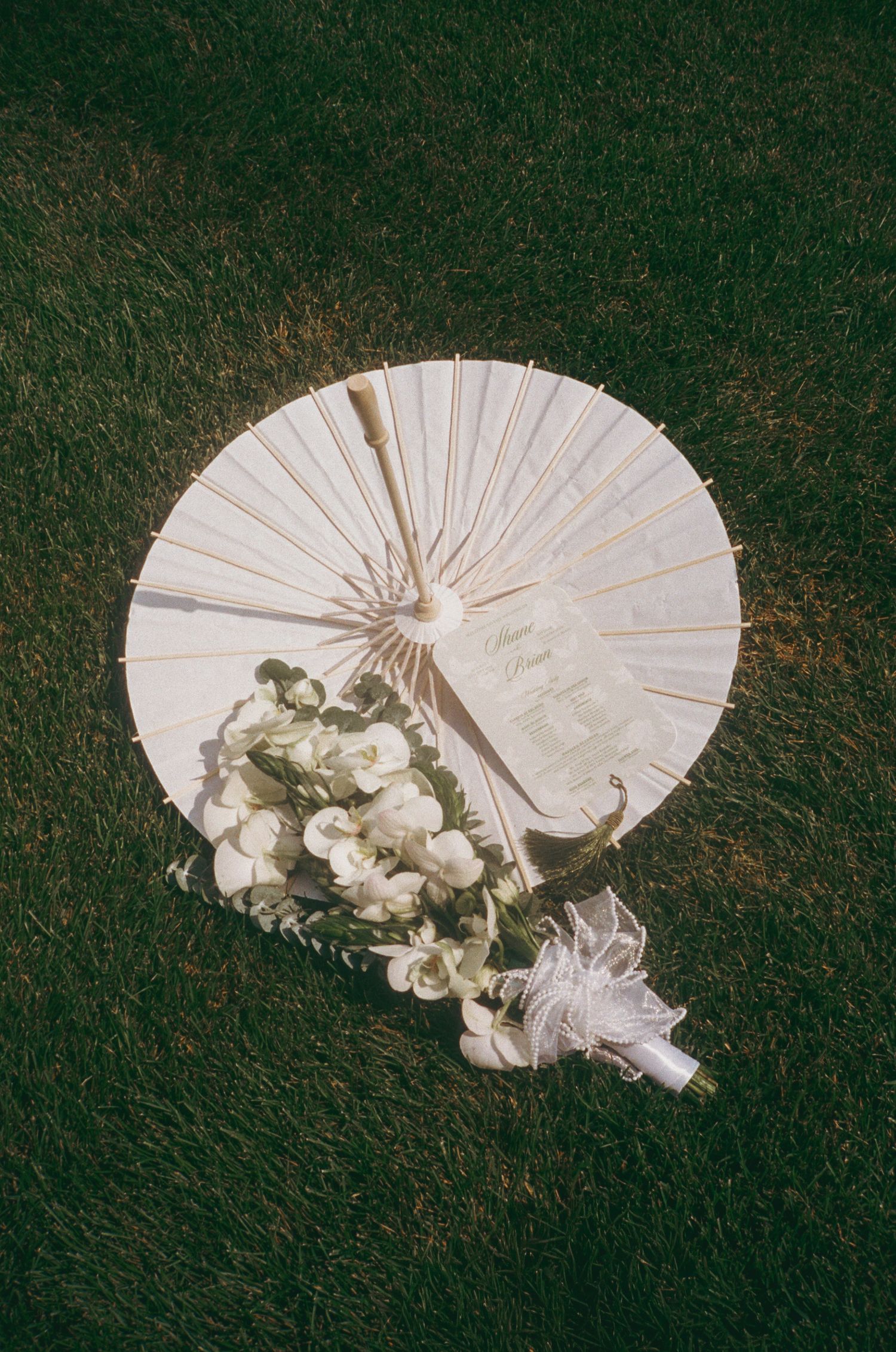 White paper parasol and bridal bouquet of white flowers resting on lush green grass.
