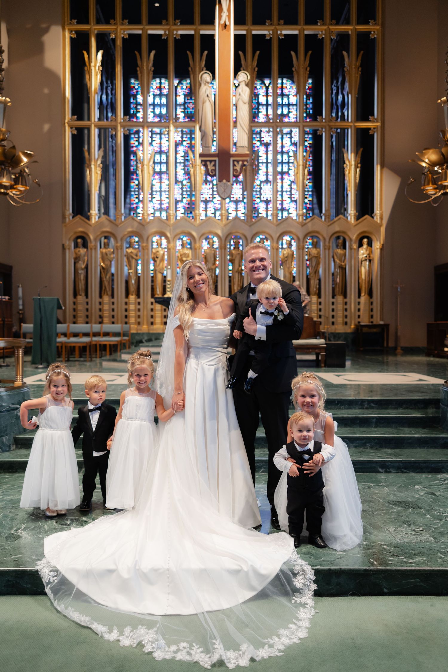 Bride and groom pose with flower girls and ring bearers at a stunning church altar with stained glass.