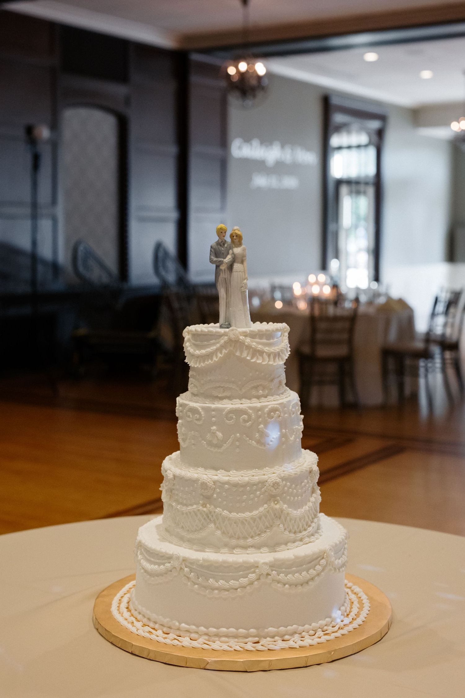 Elegant four-tier white wedding cake with lace frosting and bride and groom topper in a reception hall.