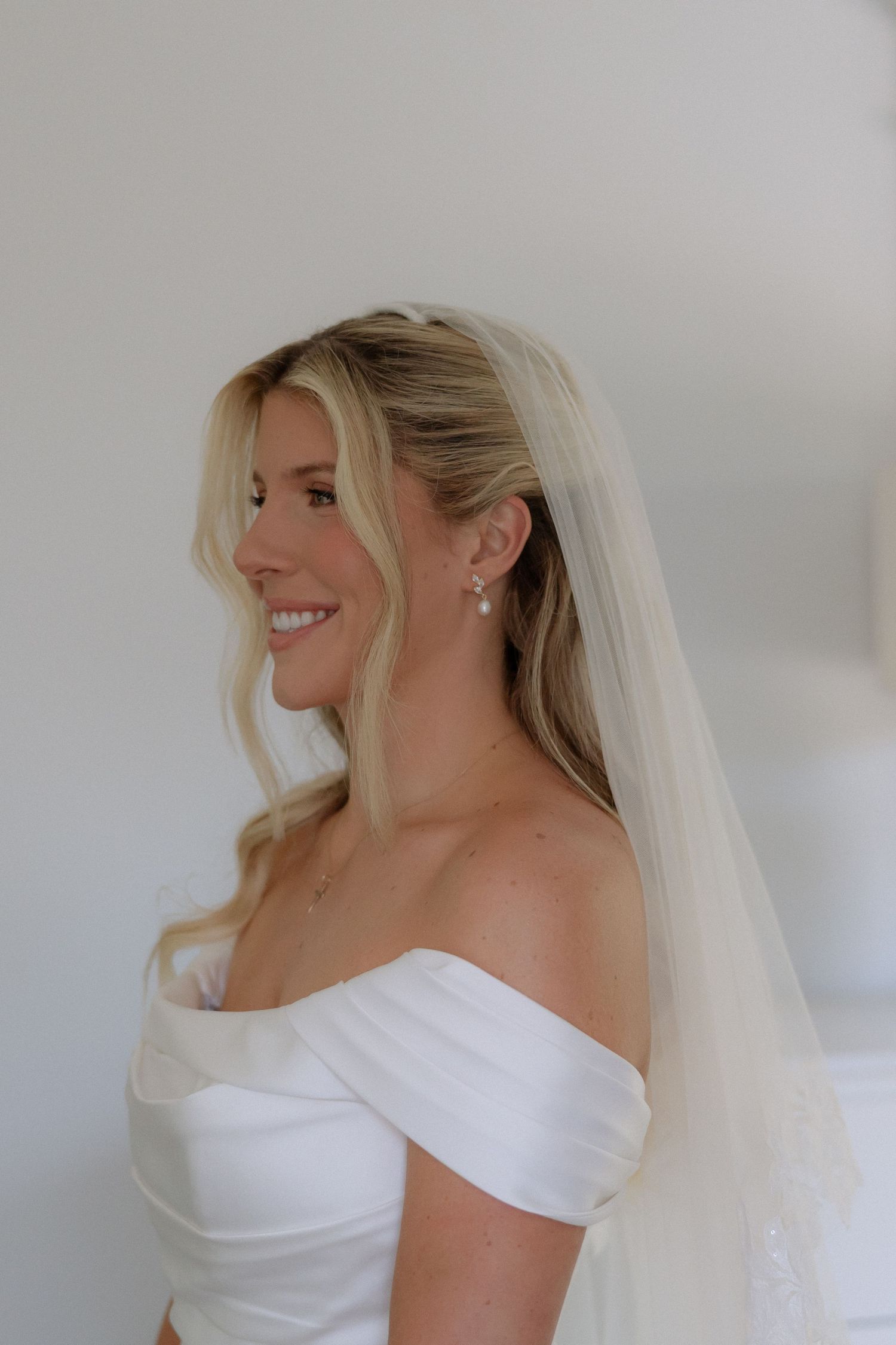 Beautiful blonde bride in an off-shoulder white gown and veil smiling softly in natural light.