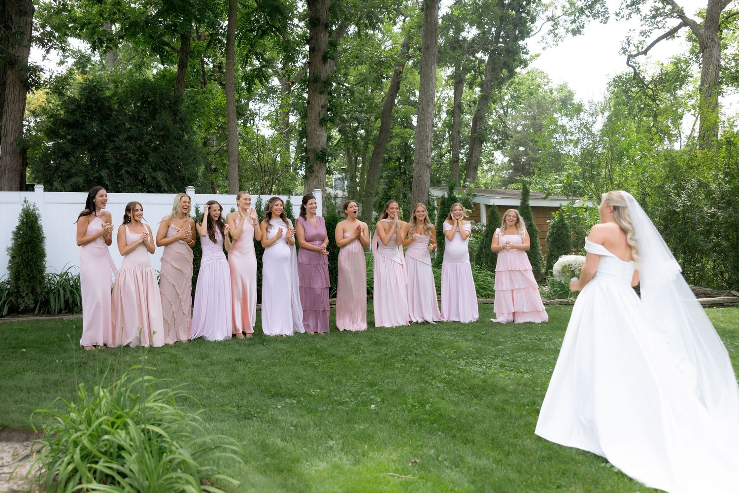 Bride reveals wedding dress to bridesmaids in pink gowns on lush garden lawn.