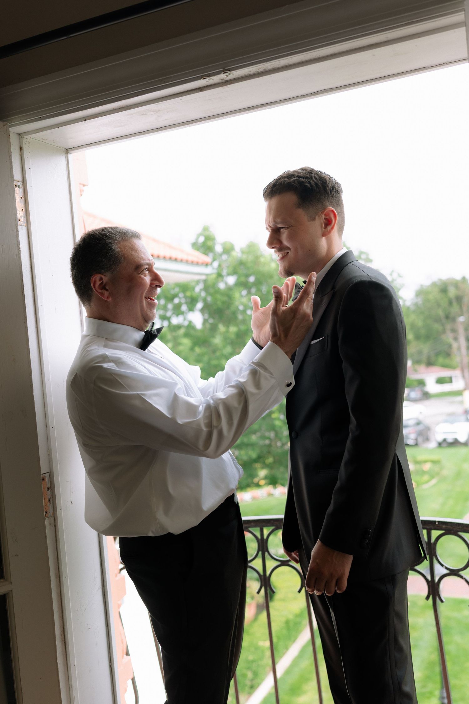 Two grooms prepare for their wedding ceremony on a balcony, one adjusting the other's bow tie.