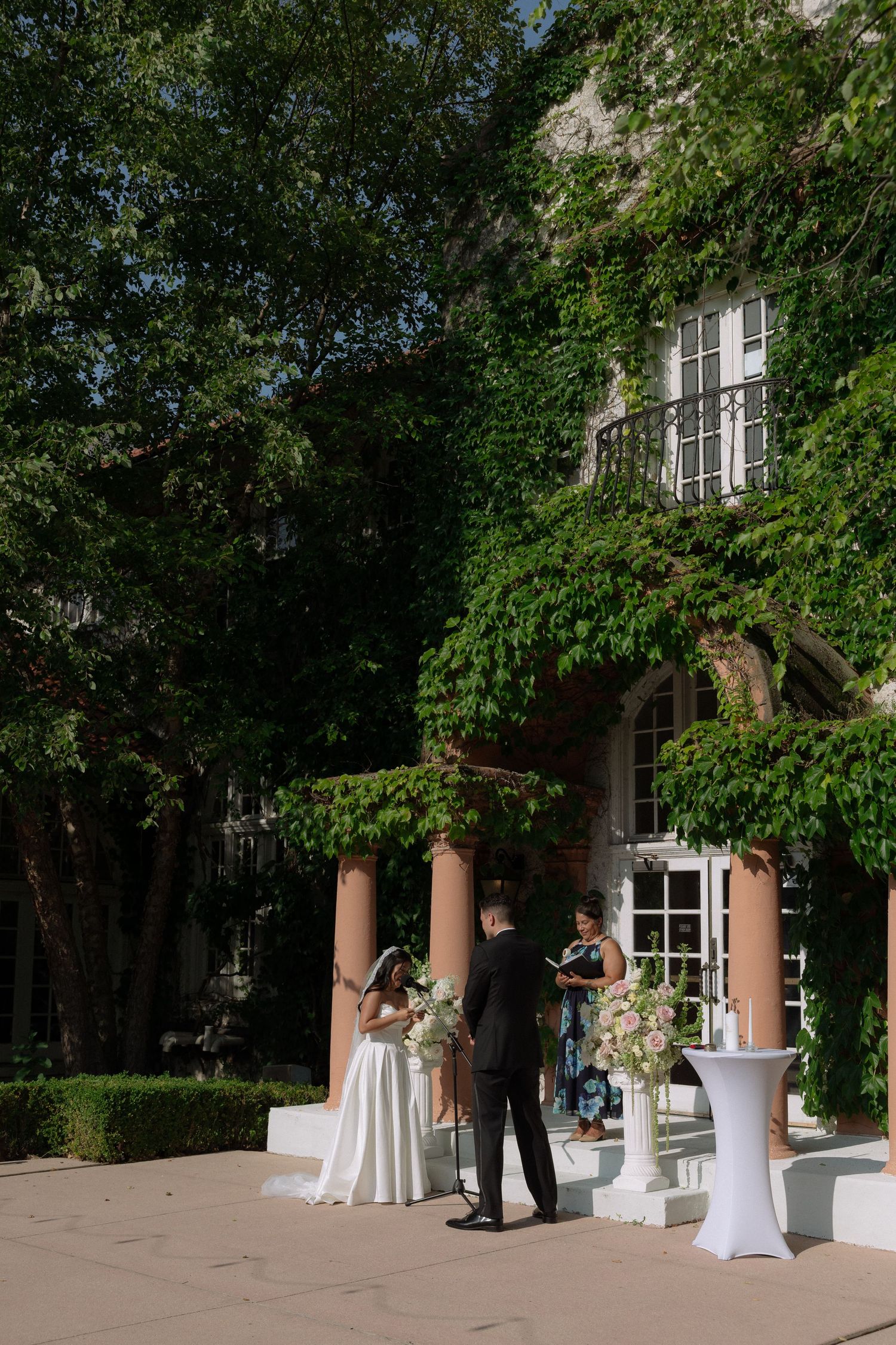 Bride and groom exchange vows outdoors at an ivy-covered stone mansion with terracotta columns and floral pedestals.