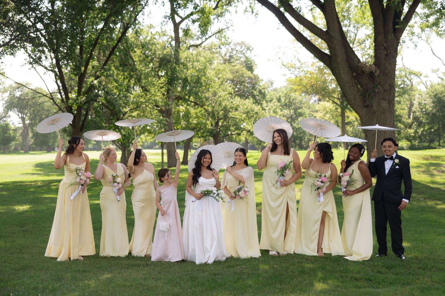 Bride with bridesmaids in yellow gowns holding white parasols under trees on sunny day.