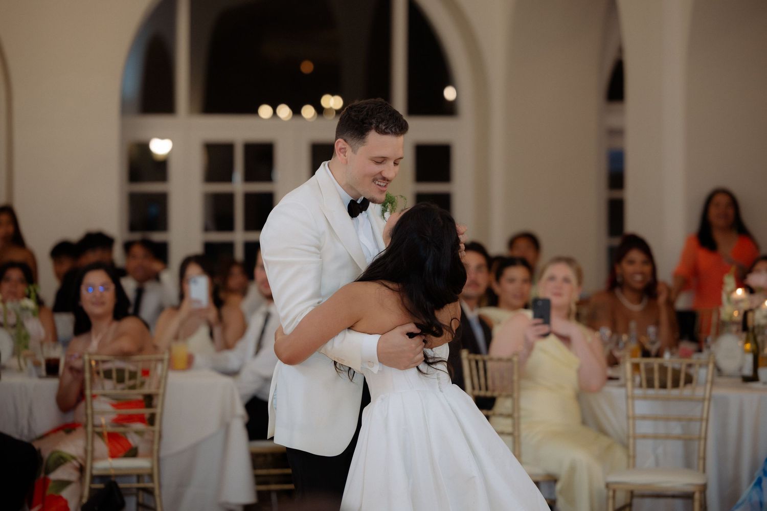 Groom in white tuxedo shares a tender first dance embrace with bride before smiling wedding reception guests.