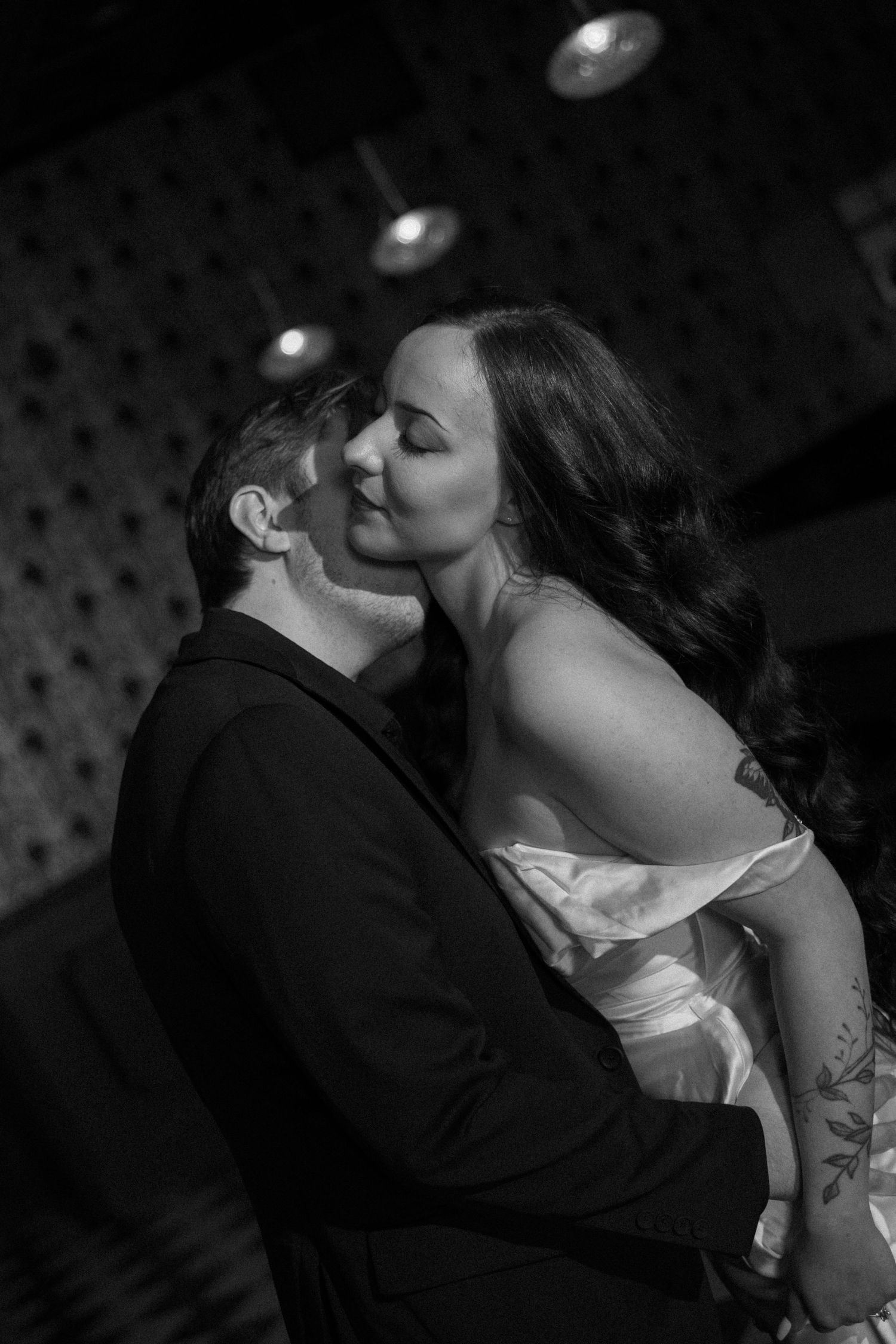 Black and white photo of couple sharing a romantic kiss, woman in white dress embracing man in dark suit.