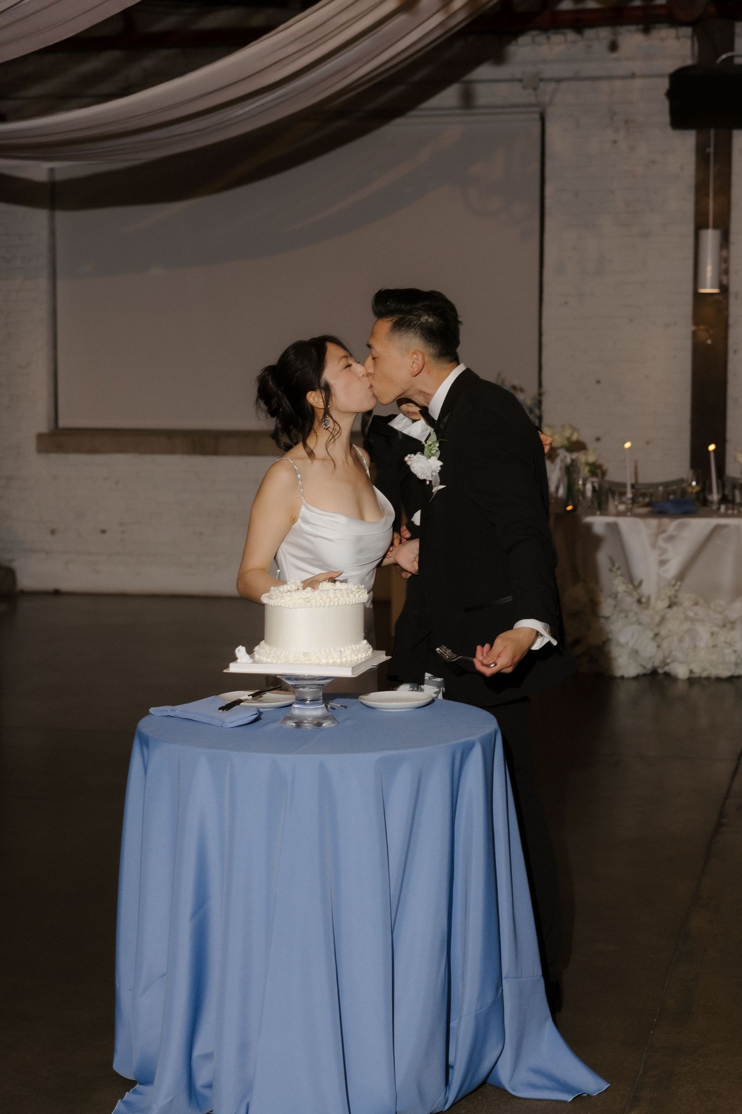 Bride and groom sharing a kiss while cutting their white wedding cake on blue tablecloth.