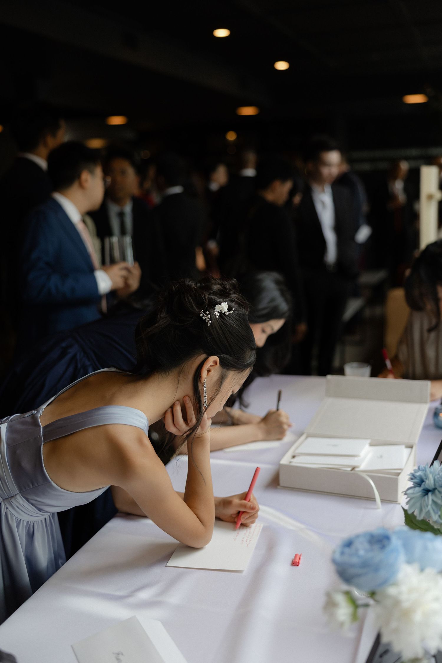 Bridesmaid in gray dress writing in wedding guest book at reception table with white flowers.