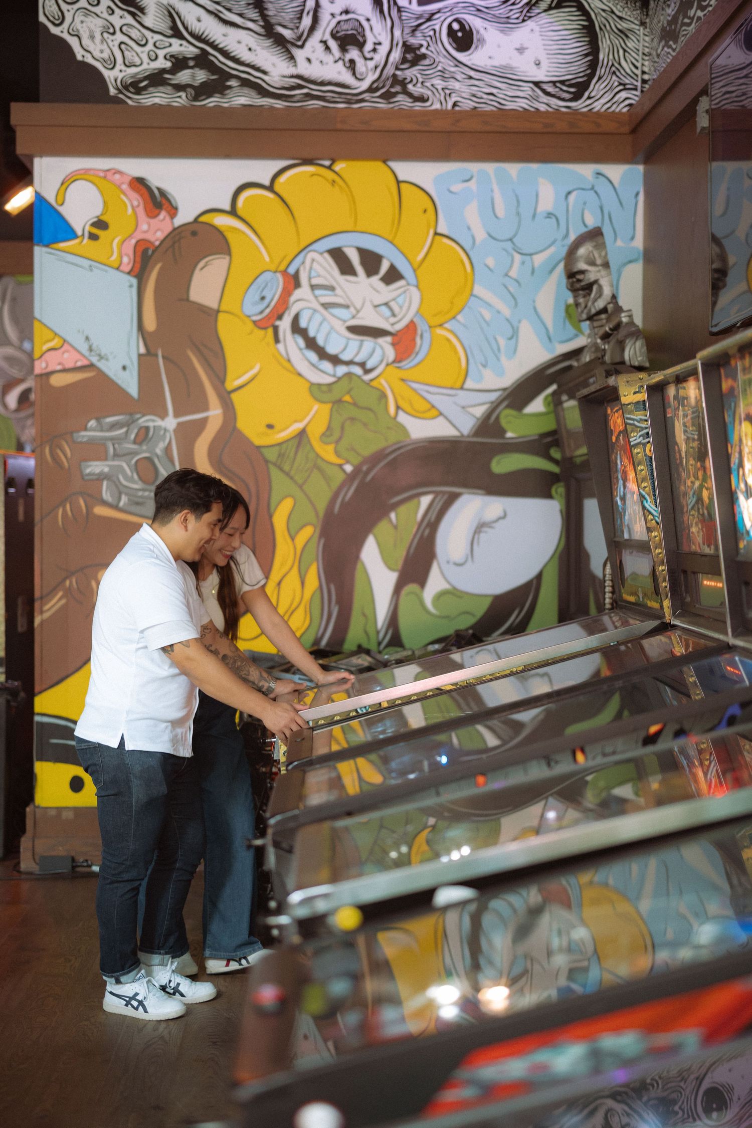 Couple playing pinball in an arcade with colorful graffiti mural featuring a cartoon sunflower on the wall.