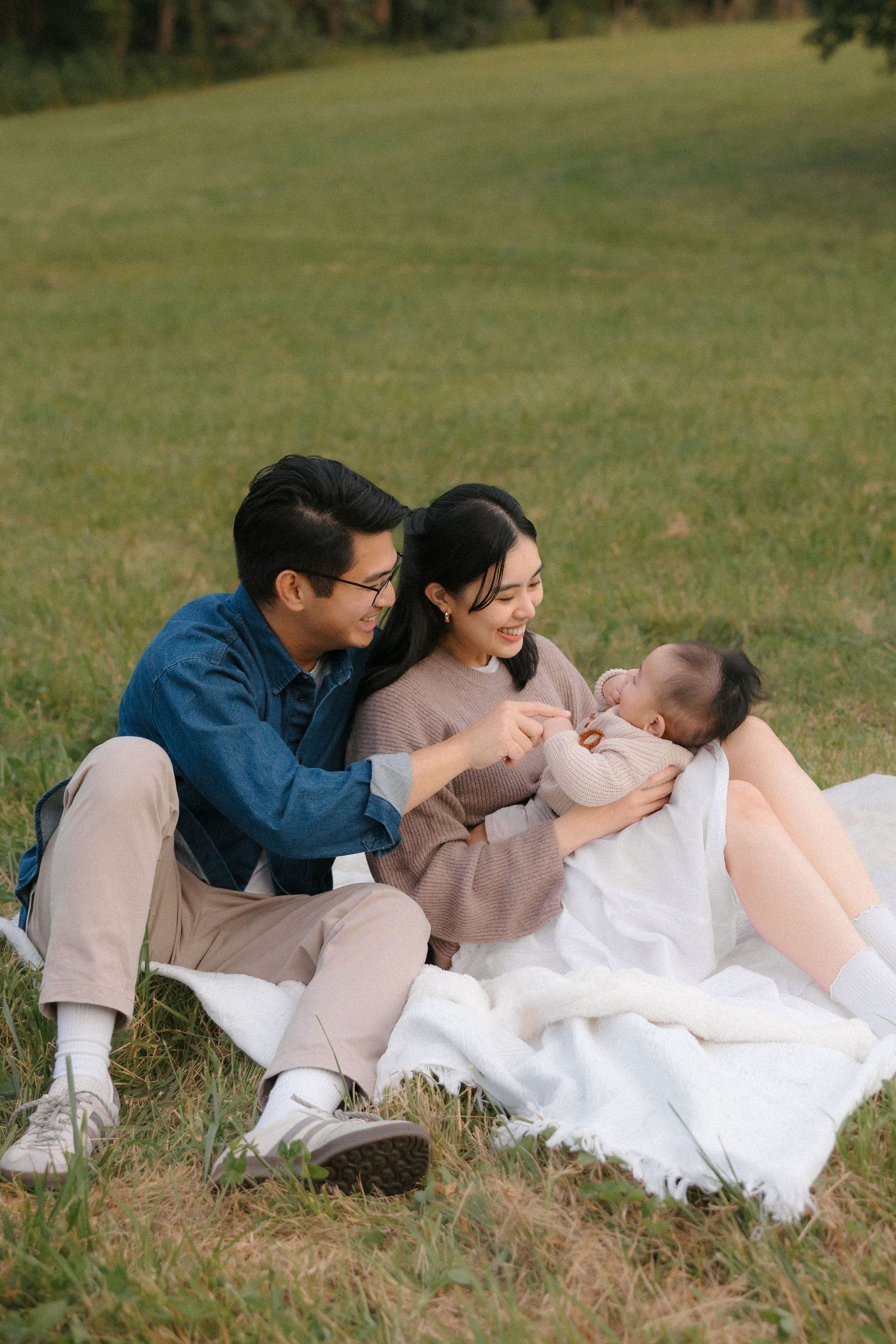 A young Asian family sits on a white blanket in a green park, smiling and playing with their baby.