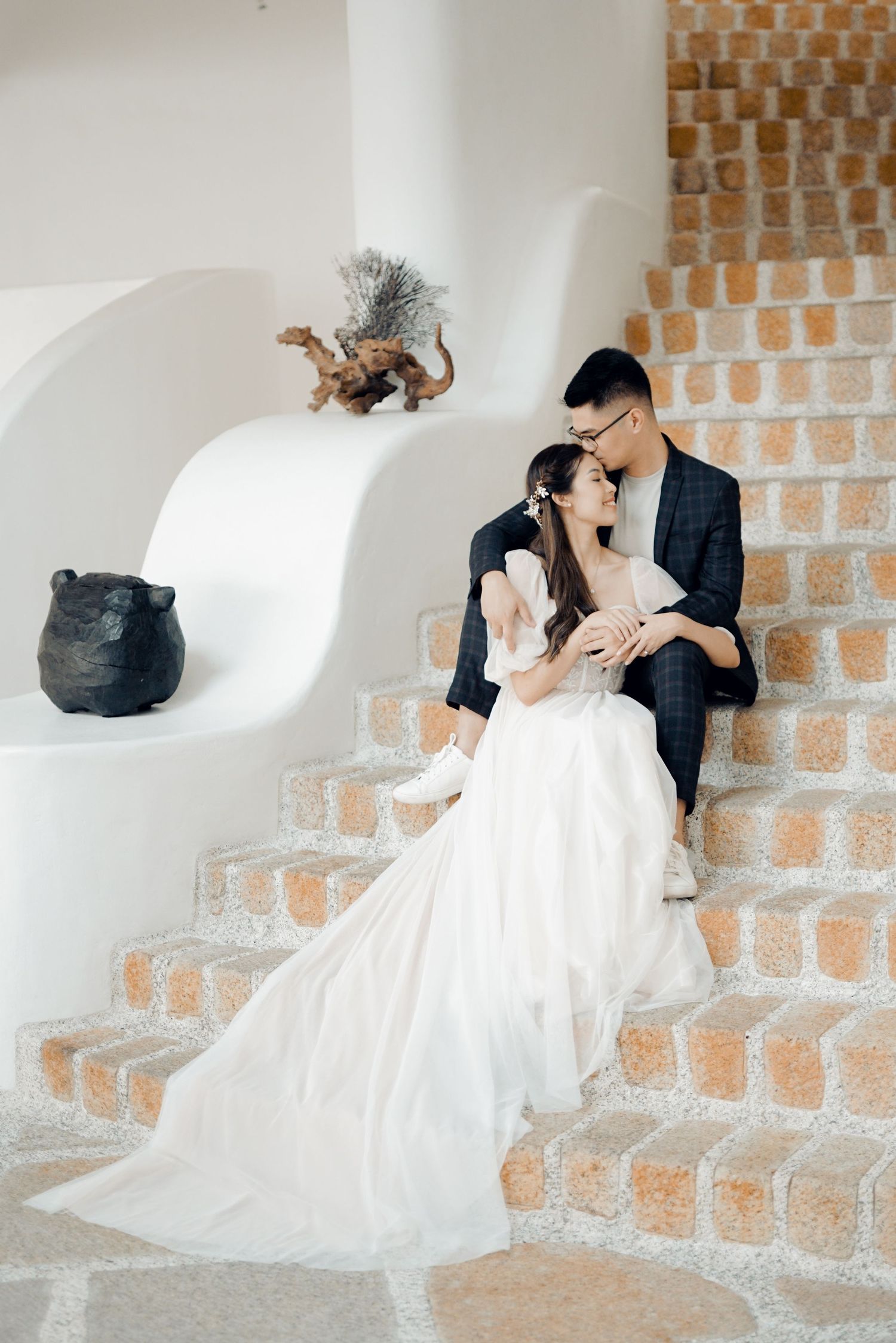 Couple in wedding attire sitting on terracotta steps, bride in white gown, groom in plaid suit, romantic pose.