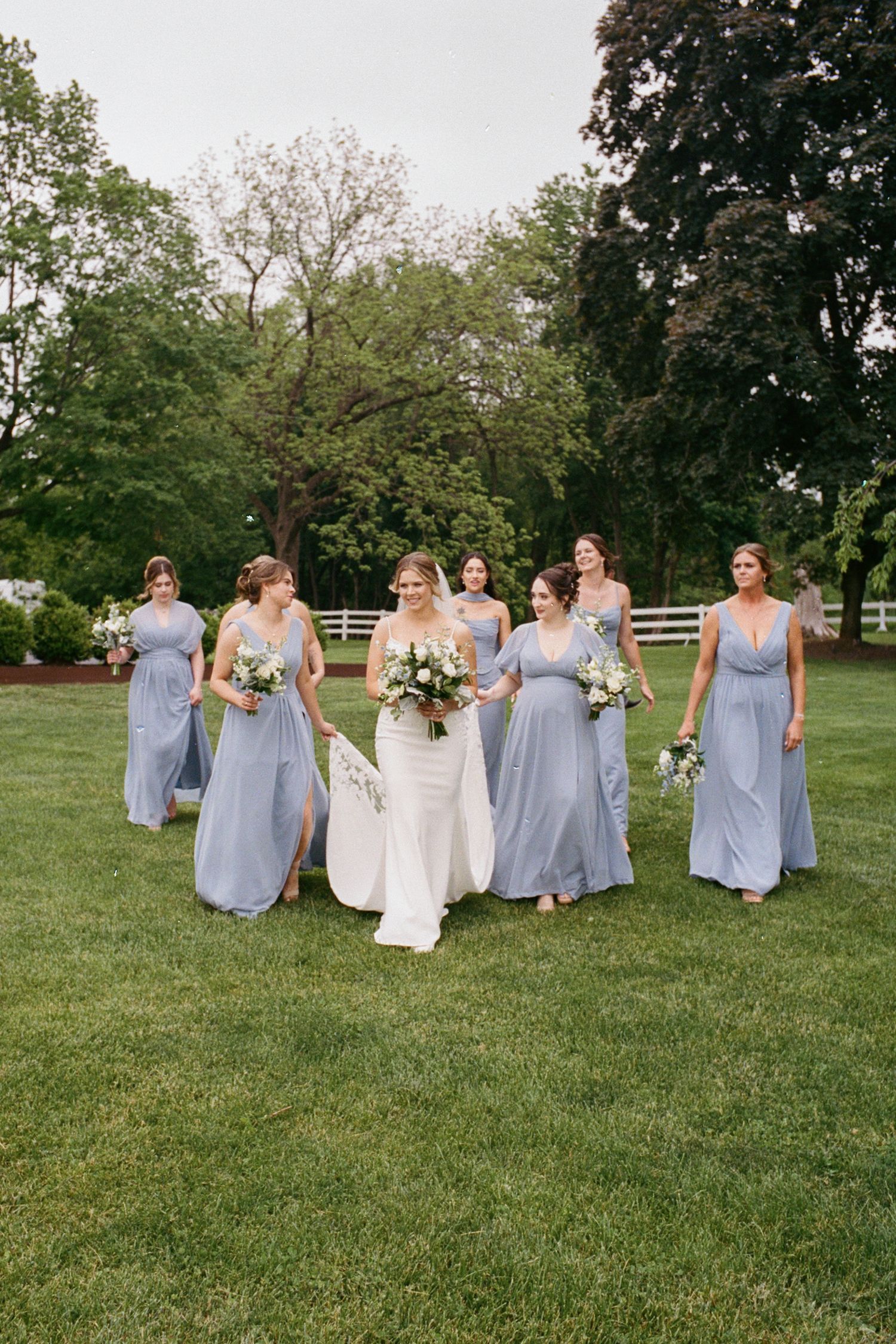 Bride in white gown walks with six bridesmaids in dusty blue dresses across a lush green lawn.
