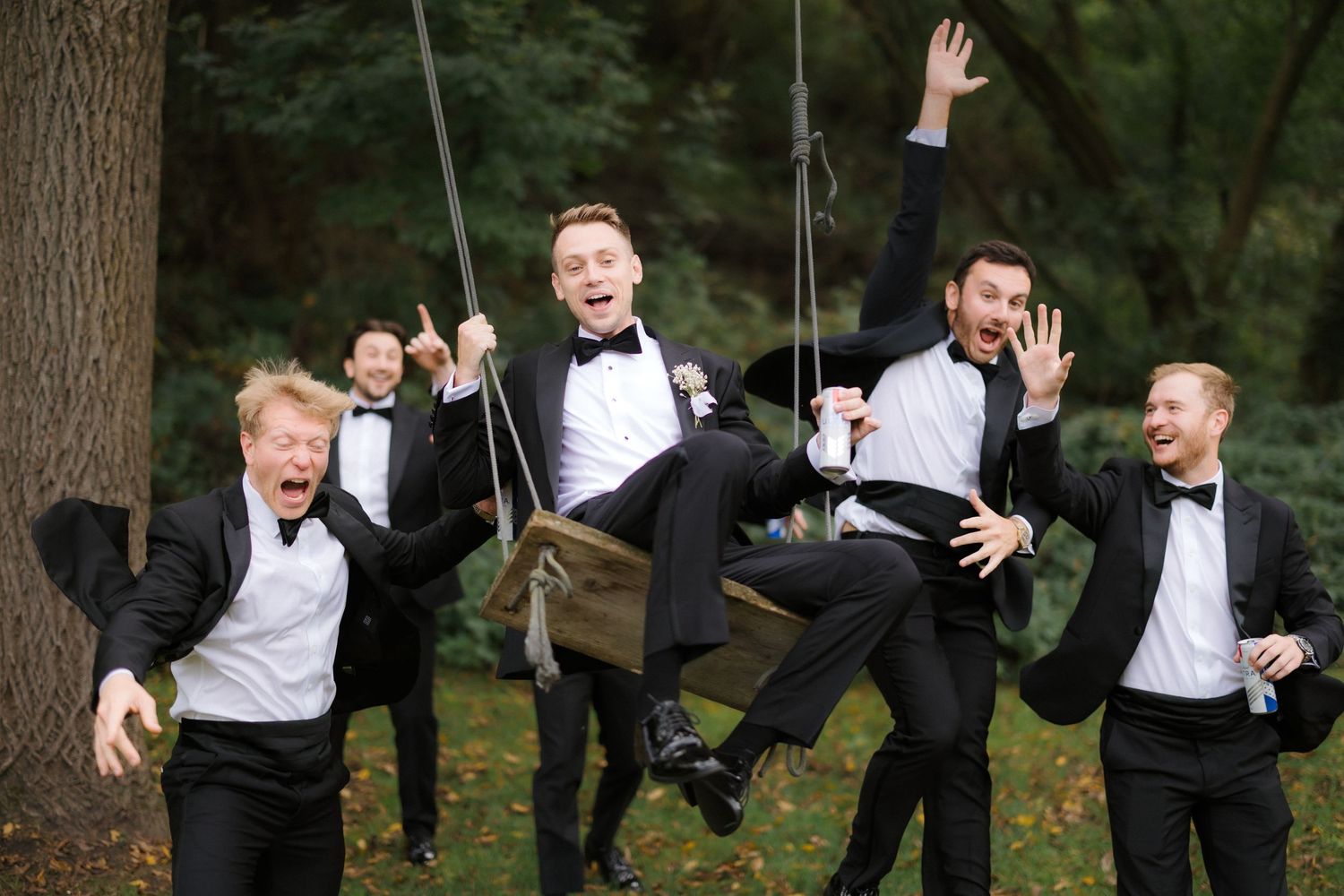 Excited groomsmen in black tuxedos celebrate with groom on swing, laughing and cheering outdoors.
