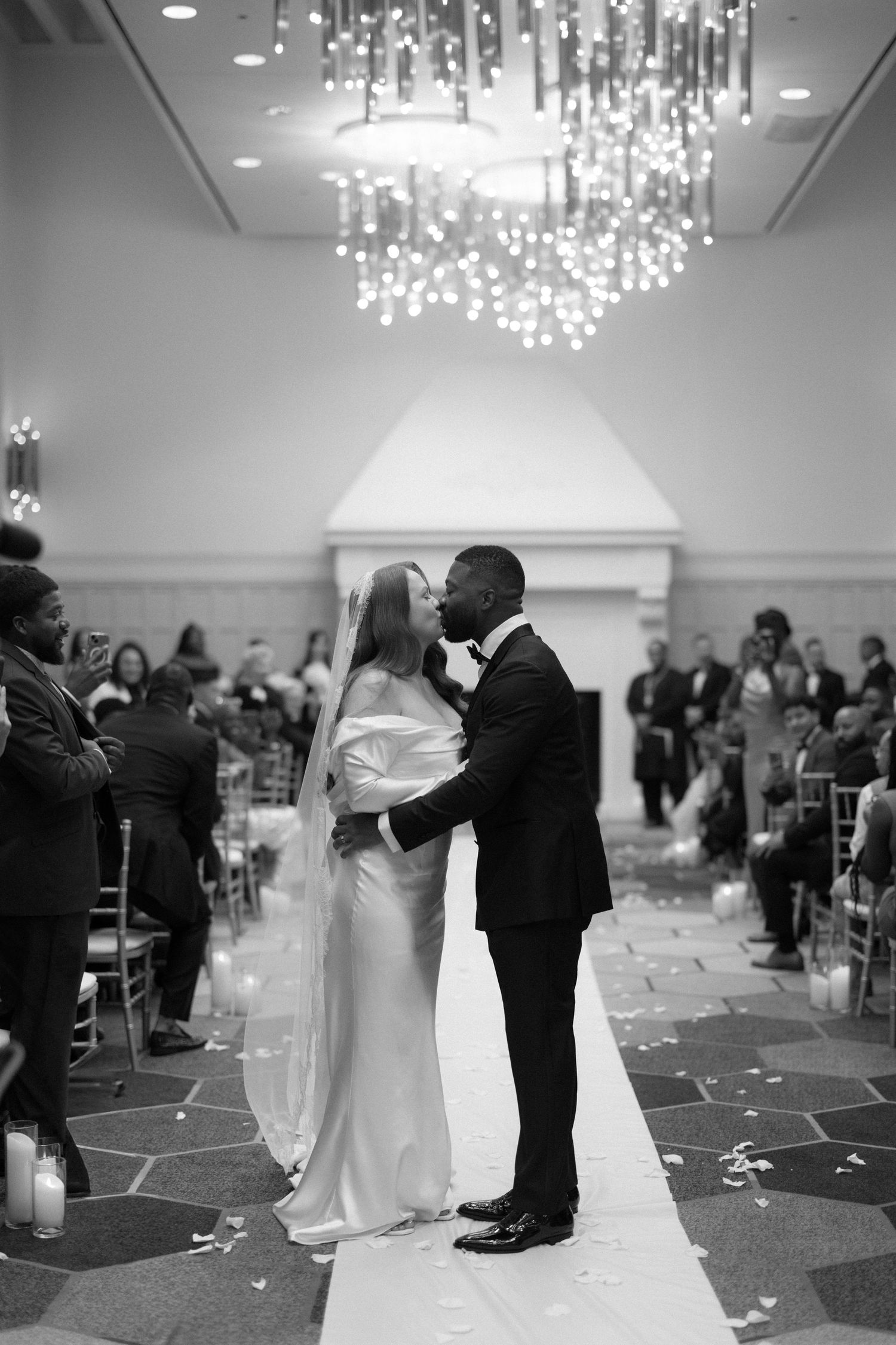 Black and white photo of a couple sharing their first kiss at a wedding ceremony in an elegant ballroom with chandelier.