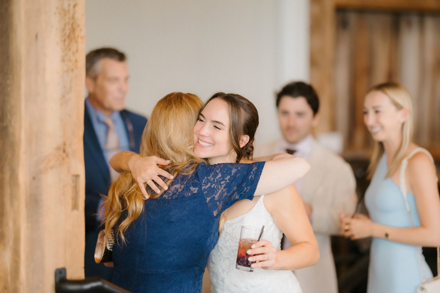 Bride in white gown sharing a warm hug with a guest in a blue lace dress at wedding reception.